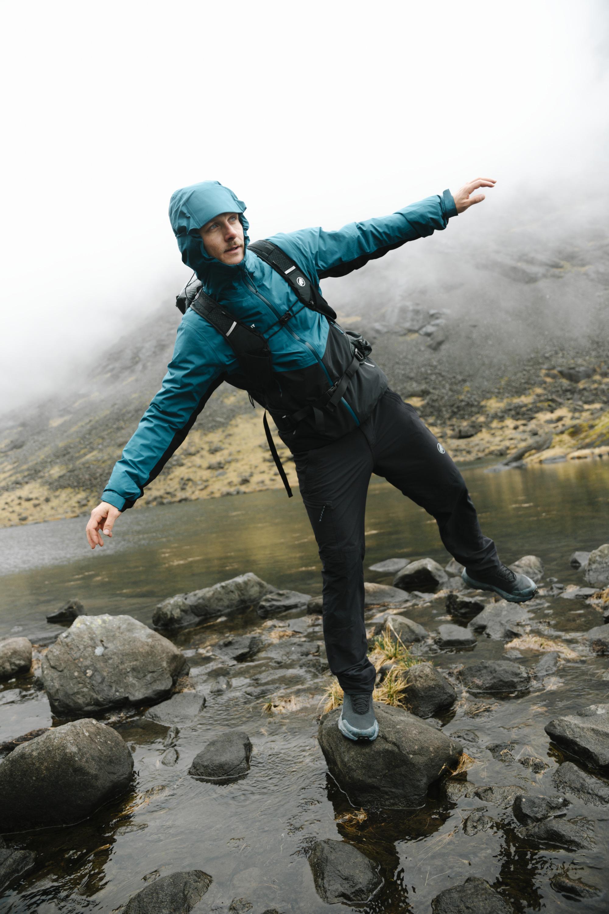 Adventurer in a teal Mammut jacket balancing on rocks beside a misty alpine lake, mountains rising in the background.