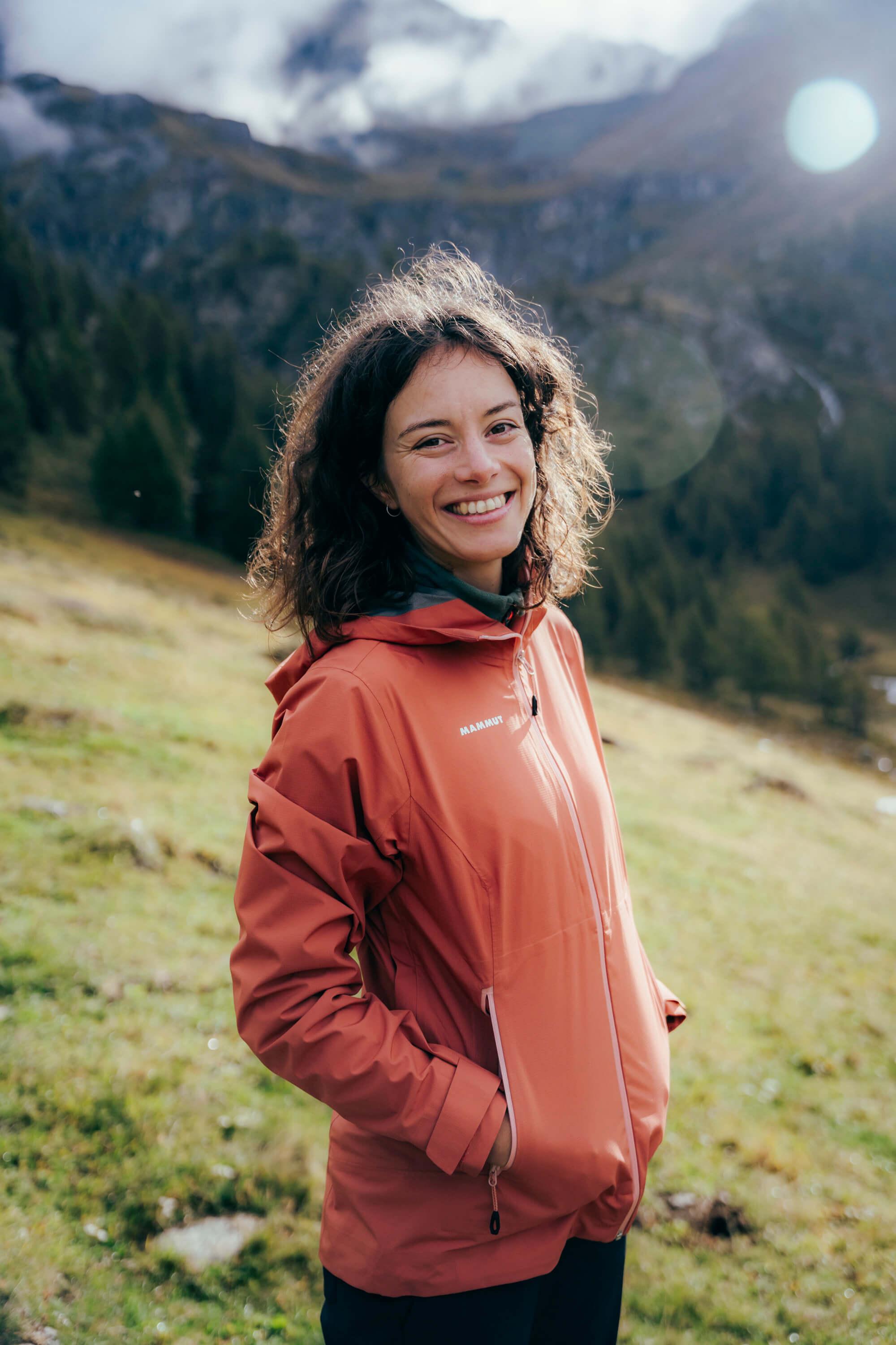 A woman in a bright orange Mammut jacket, smiling while standing in a grassy, mountainous region with a sunny, partly cloudy sky.