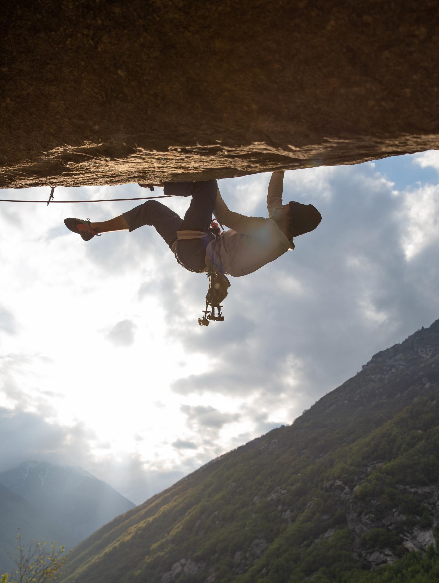 A rock climber wearing Mammut gear ascends a steep overhang with rugged mountains and a cloudy sky in the background.
