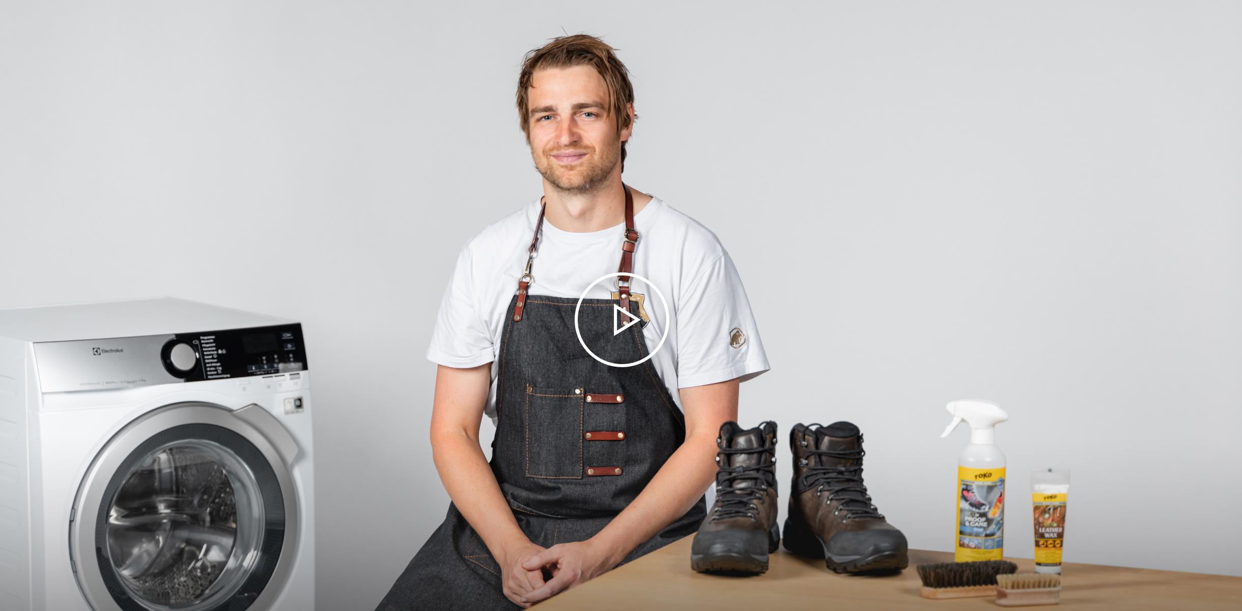 Man in Mammut apron sitting by a washing machine with mountaineering boots and cleaning supplies on a table, posing for a photo.