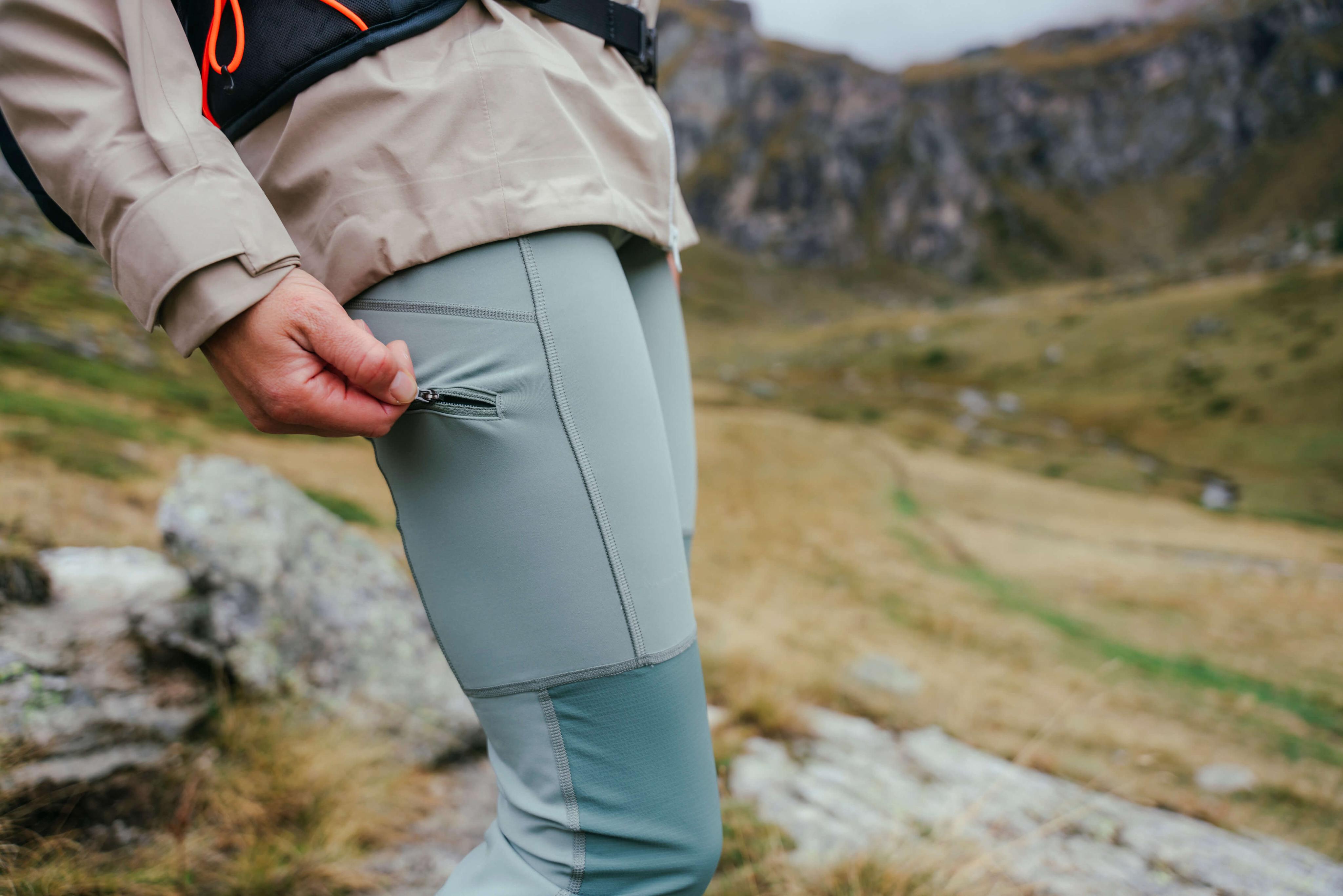 Person in Mammut outdoor gear unzipping a pocket on their hiking pants while trekking through a rugged mountain landscape.