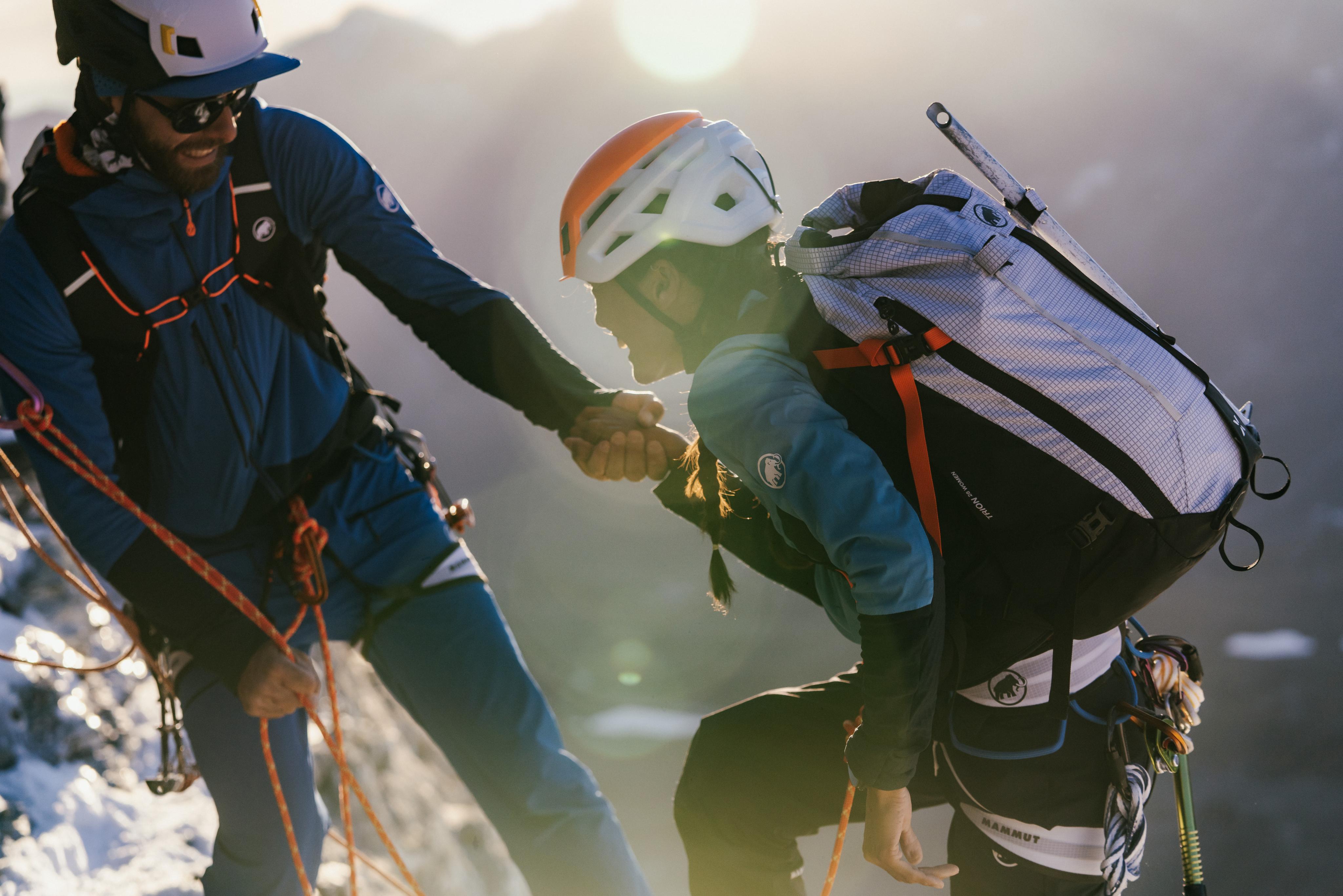 Two Mammut-clad climbers in helmets assist each other on a snowy mountain peak with the sun shining brightly in the background.