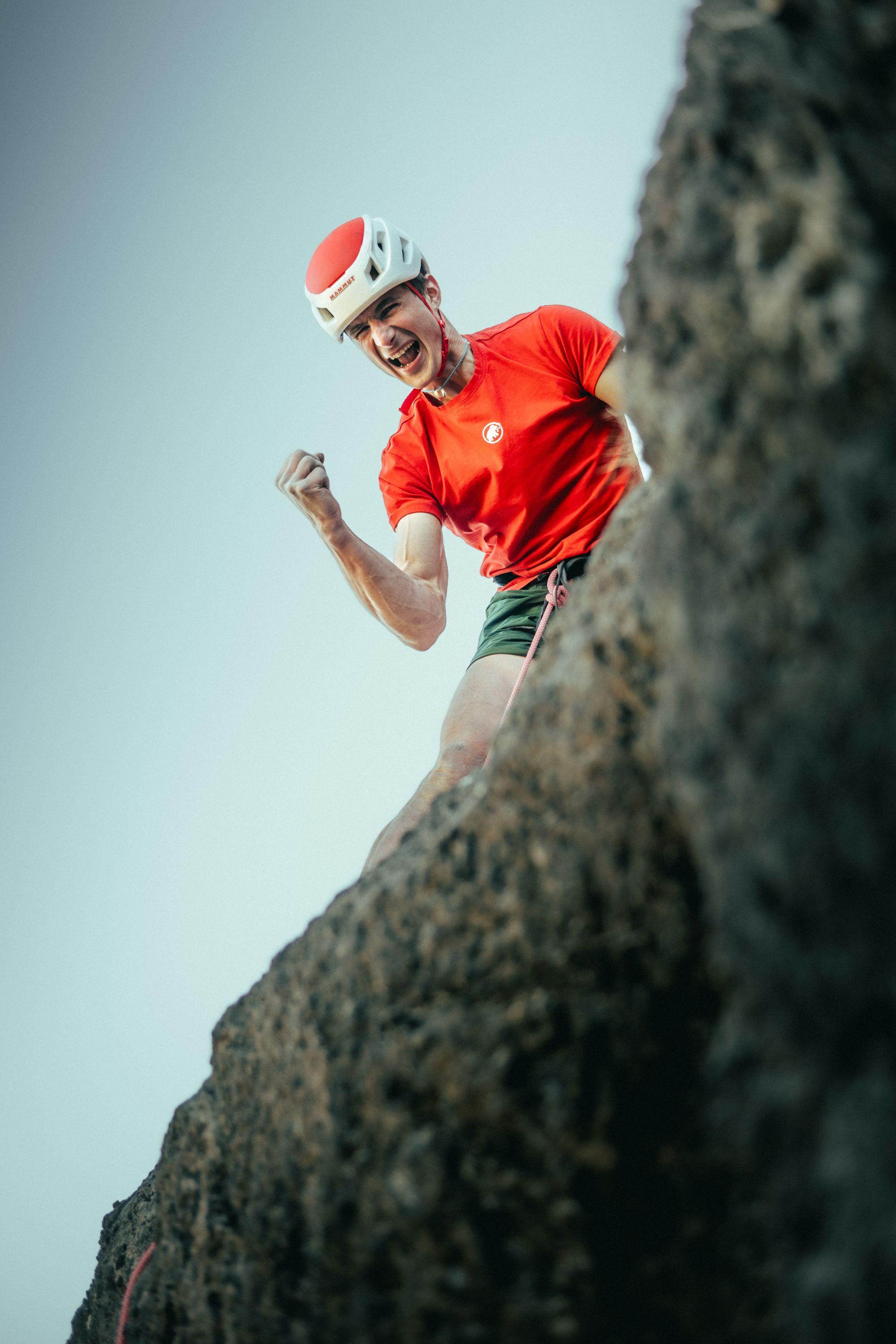 A climber in a red shirt and helmet, wearing Mammut gear, celebrates triumphantly atop a rocky cliff after a successful ascent.