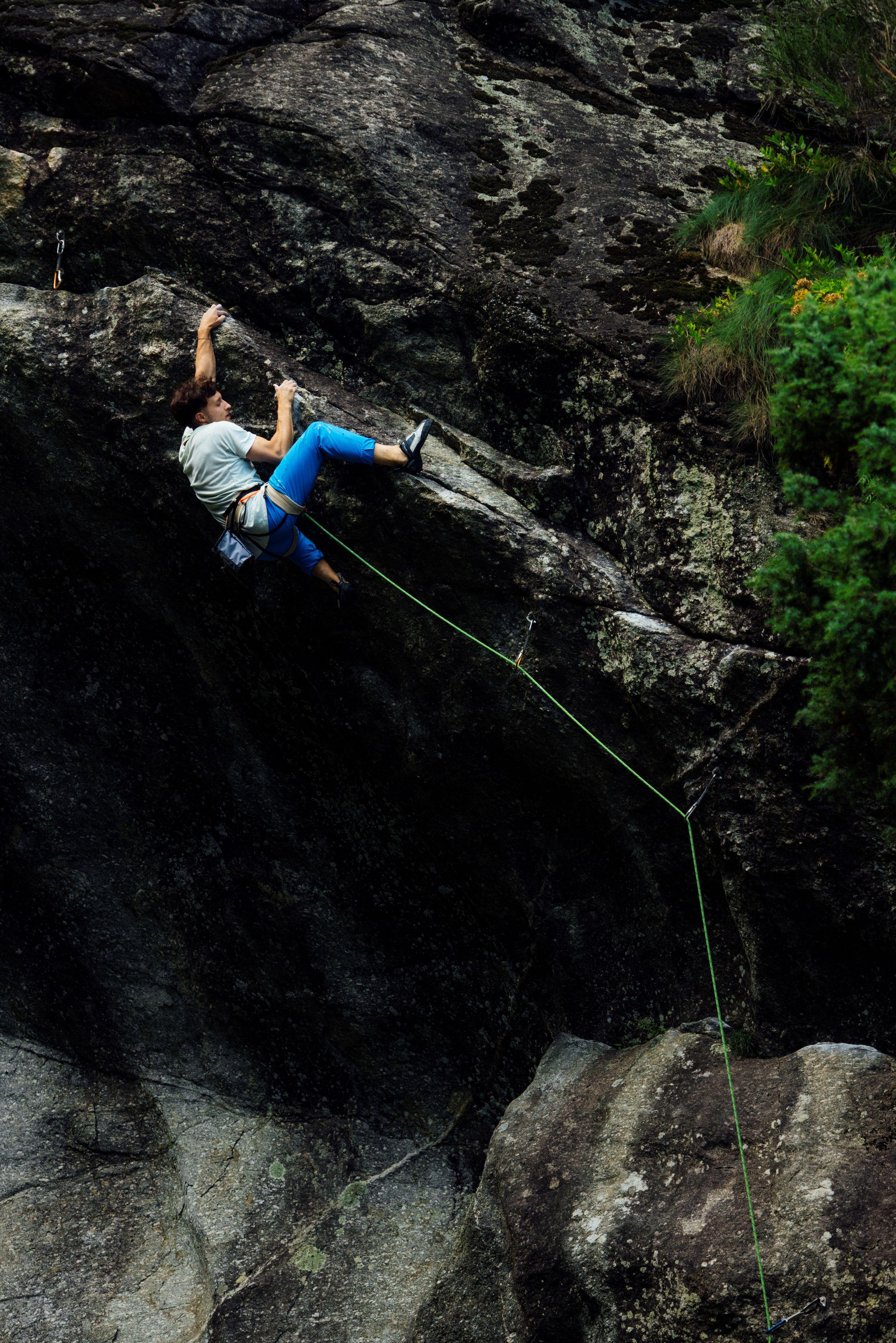 A climber in a Mammut harness adjusts their gear near a rock wall, wearing a sleeveless top and focused on the task.