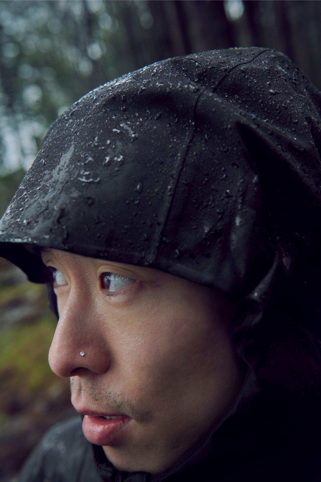 Close-up of a person wearing a Mammut rain jacket with hood, covered in raindrops, outdoors in a forest.
