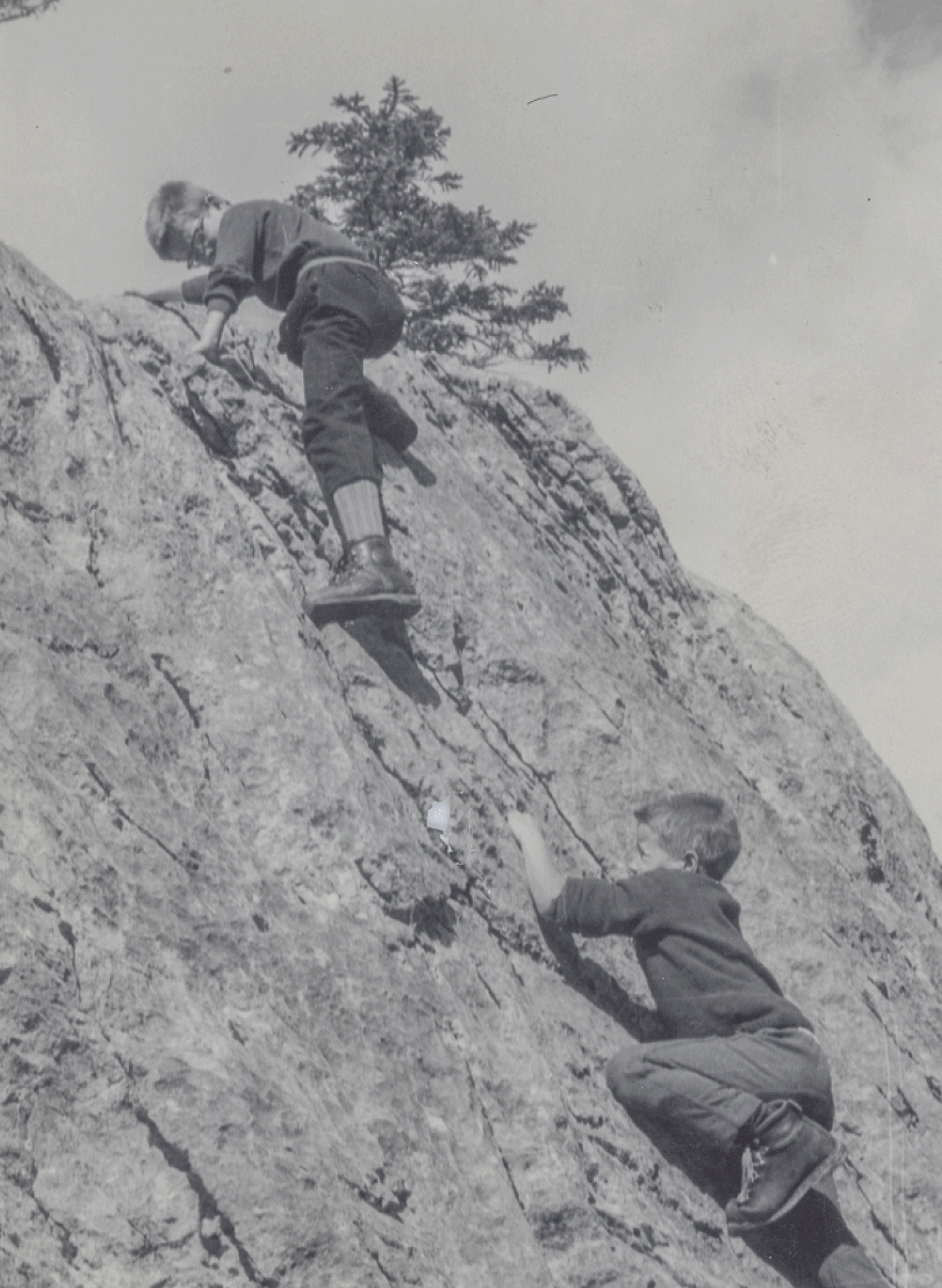 Two children wearing Mammut climbing gear scale a large rock formation, with one child higher up and the other following closely behind. Trees and blue sky provide a scenic background.