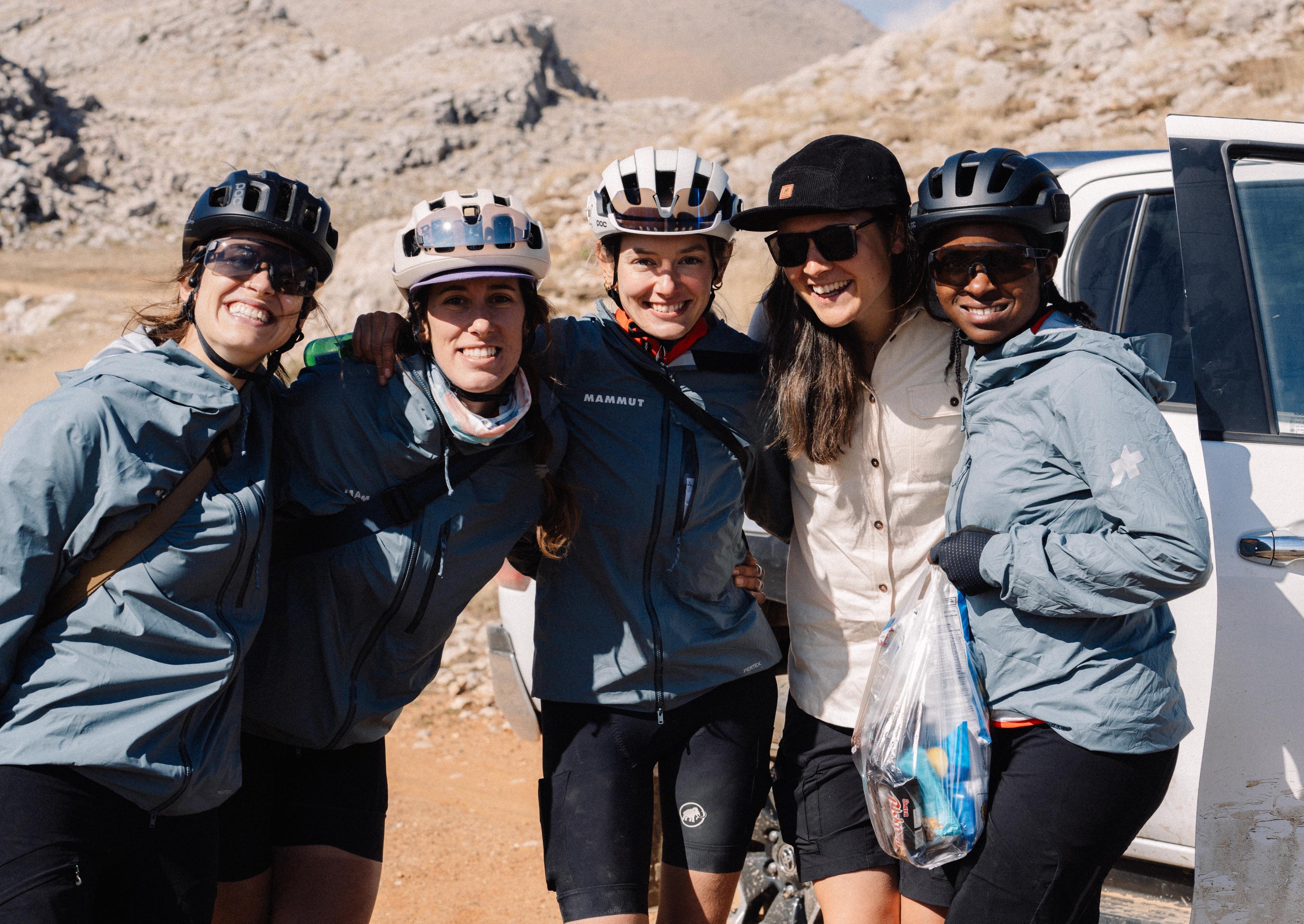 Five Mammut-clad cyclists in helmets and jackets smile and pose together in a rugged, rocky landscape.