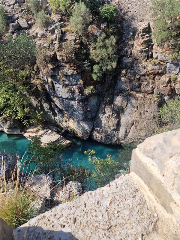 Cliffside view overlooking a blue river cutting through rocky terrain, with lush greenery and a hiker wearing Mammut gear in the foreground.