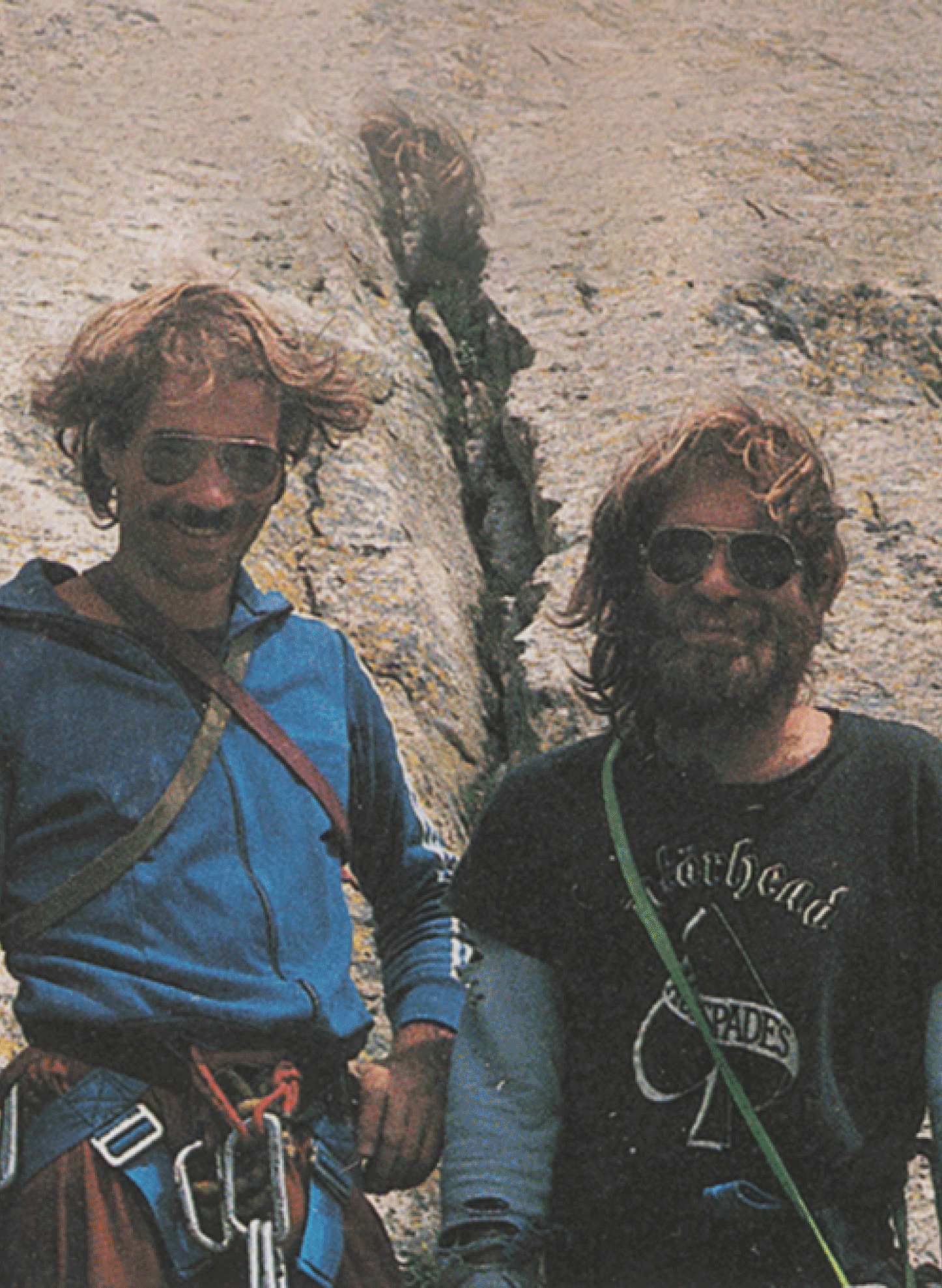 Two climbers with windblown hair standing against a rugged rocky backdrop, wearing Mammut climbing gear and sunglasses.