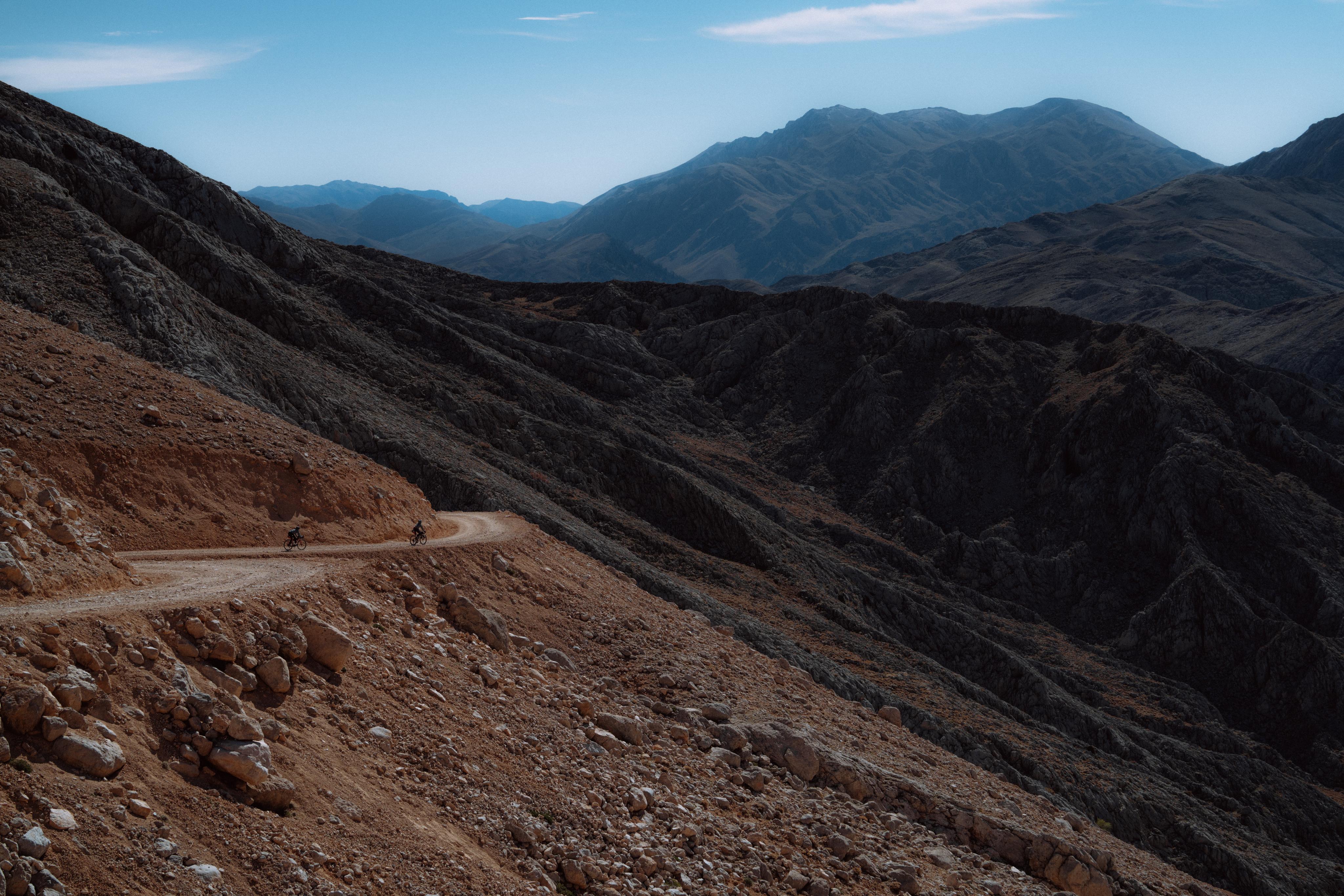 A cyclist rides along a rugged mountain path under a clear blue sky, donning Mammut gear, with distant snow-capped peaks in the background.