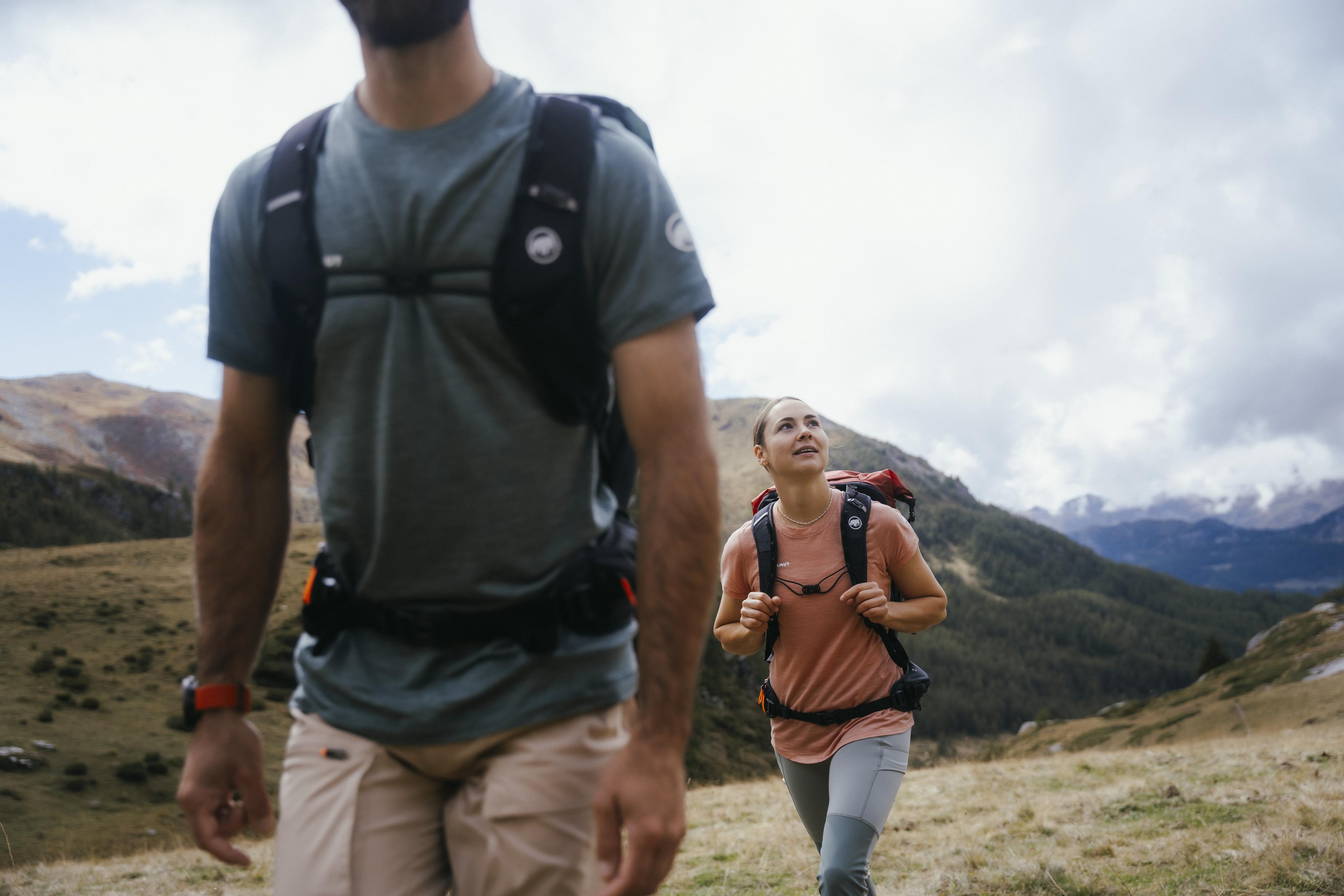 Two hikers with Mammut backpacks traverse a scenic mountain trail under a partly cloudy sky.