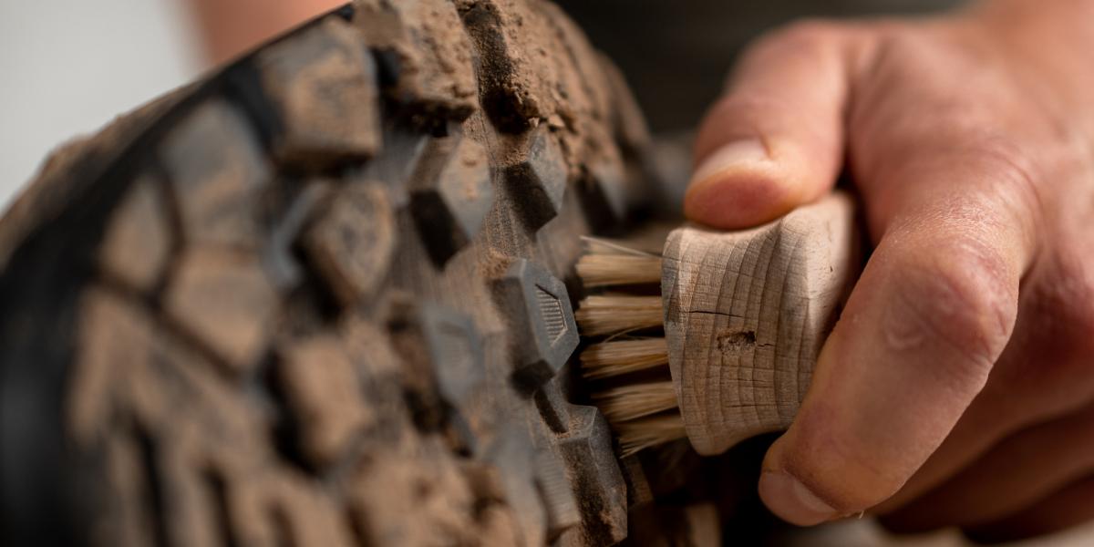 A hand uses a brush to clean dirt from the tread of a muddy Mammut hiking shoe.