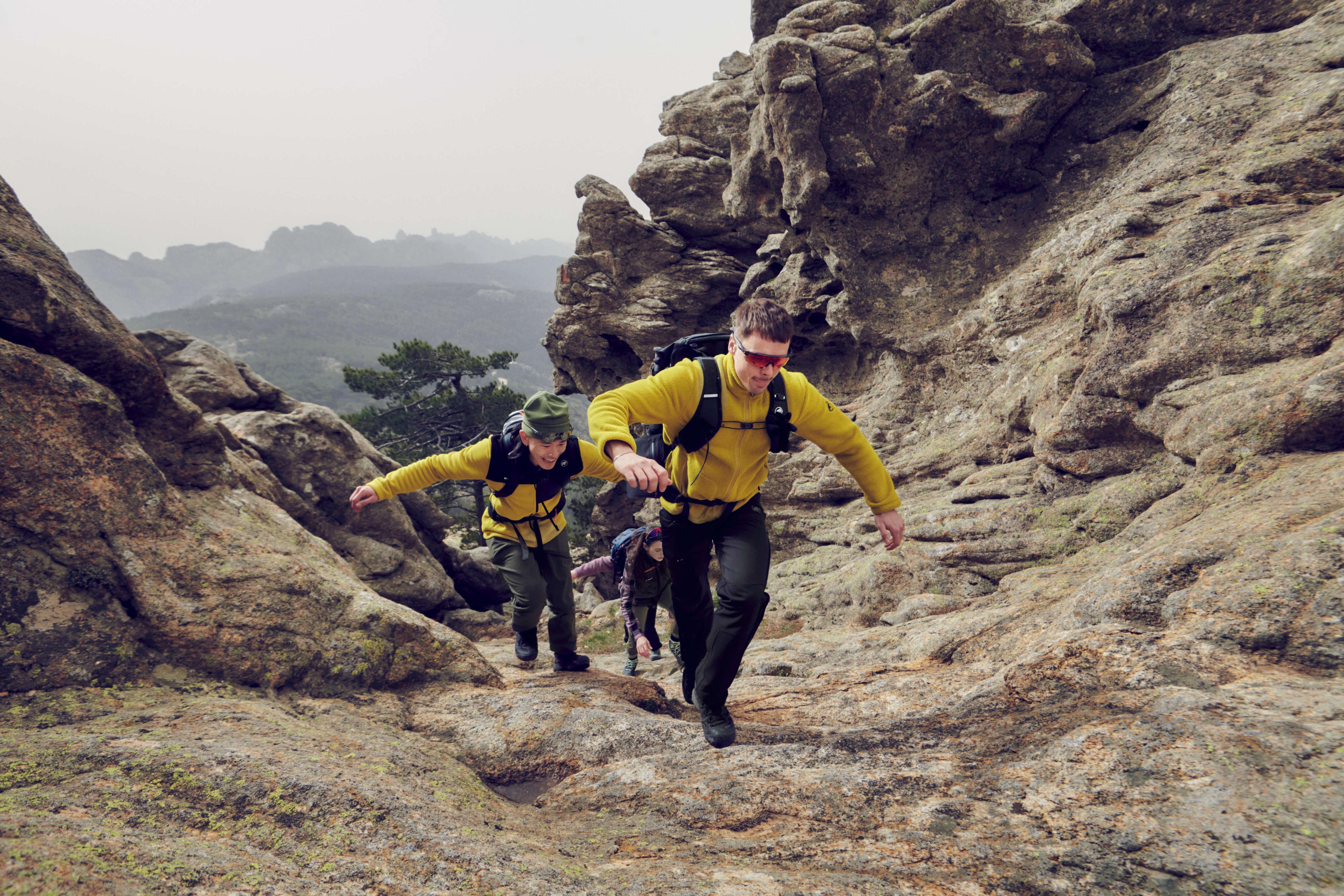 Three hikers in yellow Mammut jackets climb up a rocky mountain trail surrounded by rugged, steep alpine terrain.