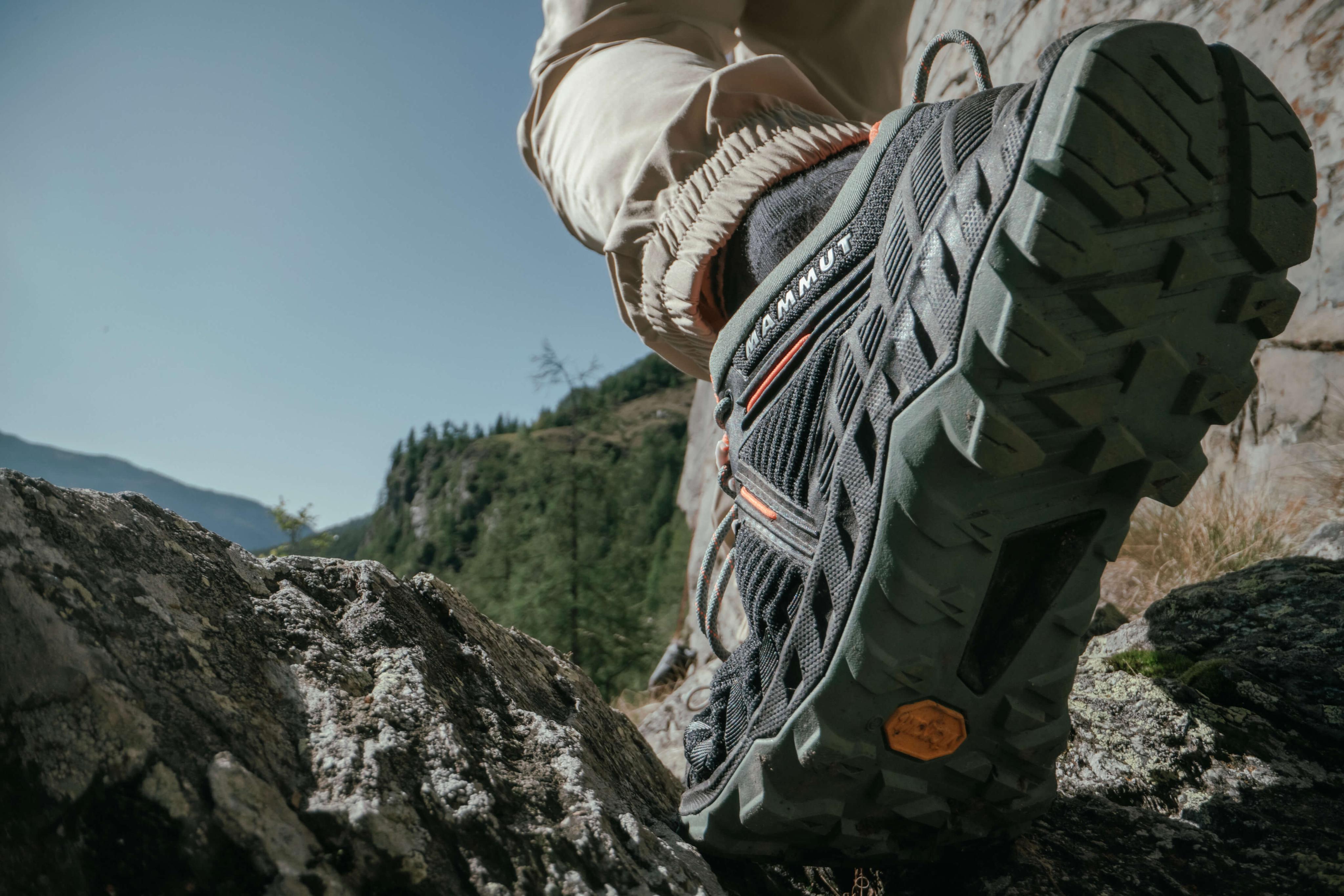 Close-up of a hiker's Mammut hiking shoe stepping on rocky terrain, with a stunning mountain landscape in the background under a clear sky.