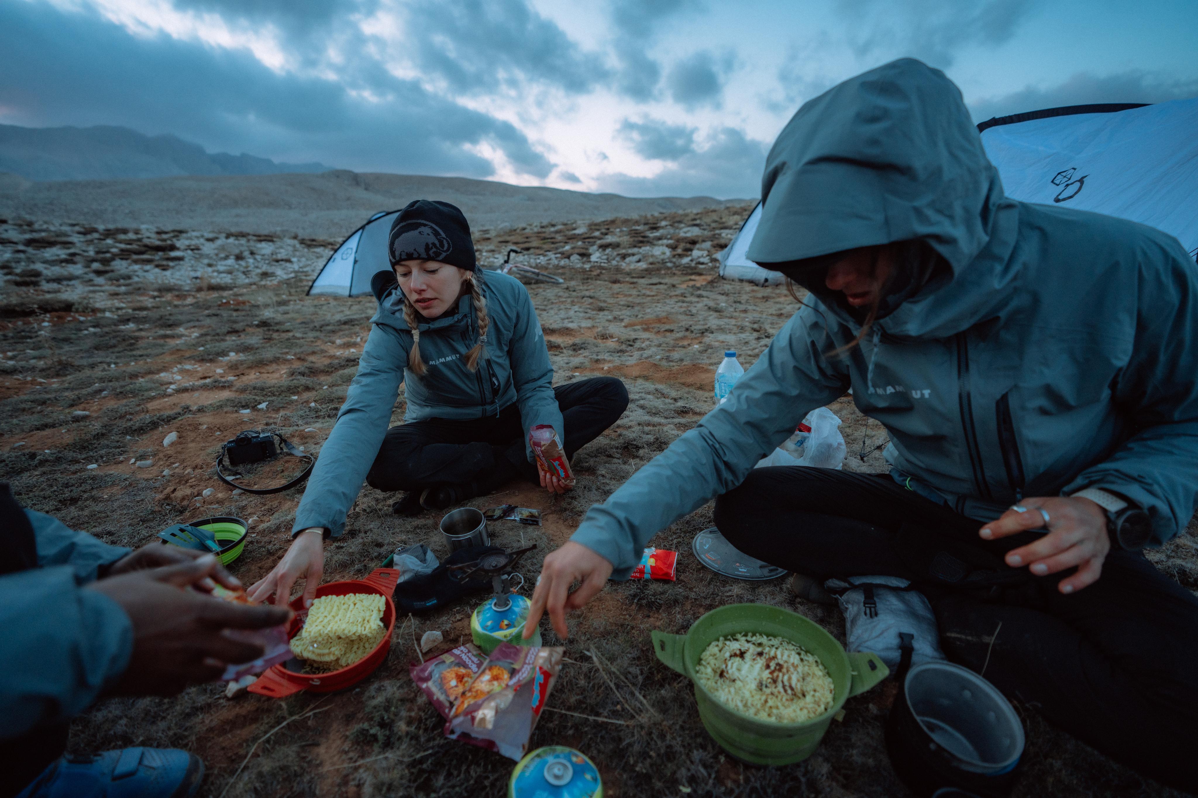 Two adventurers in Mammut outdoor gear cooking by their tents on rugged rocky terrain under a dramatic cloudy sky.