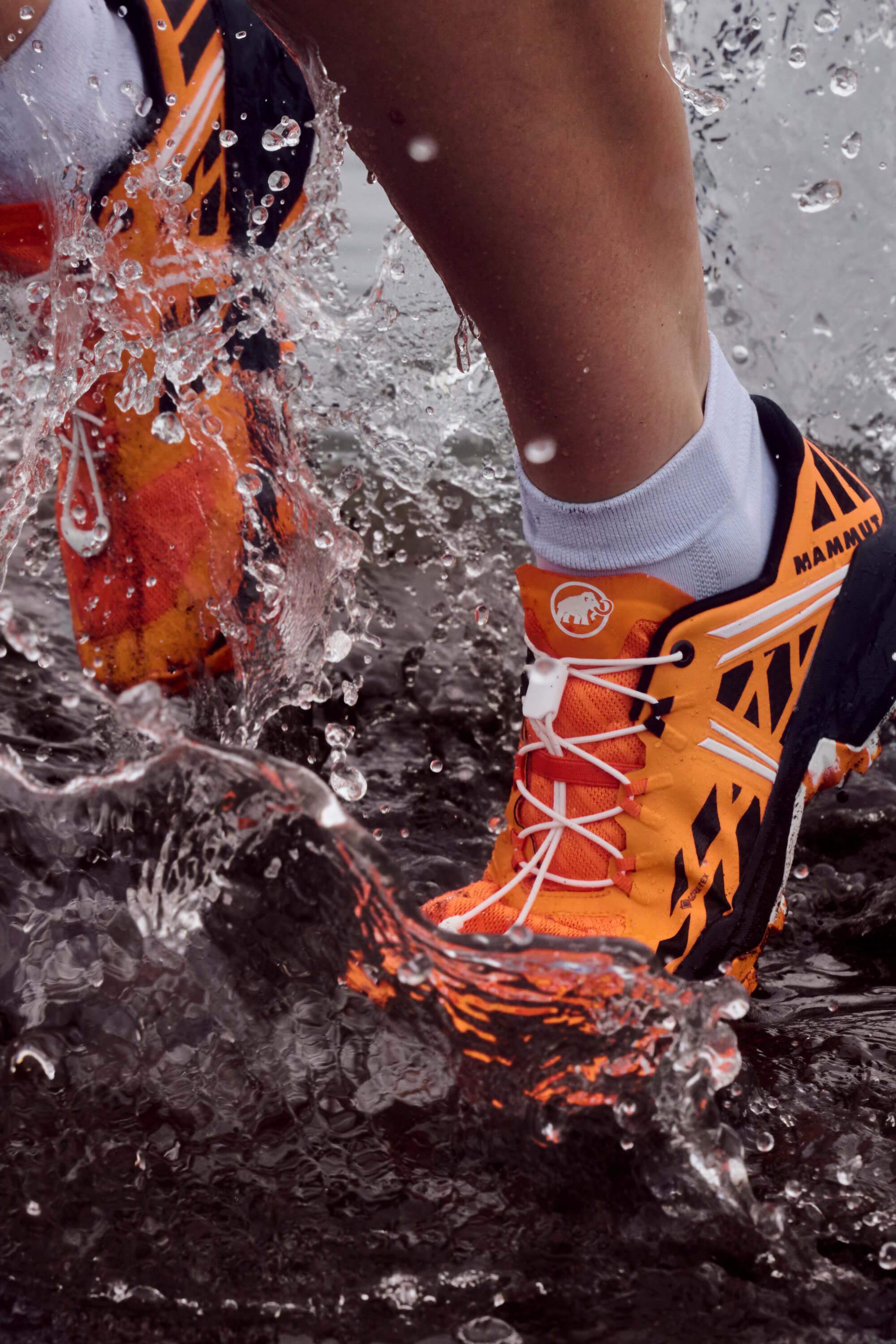 Close-up of a runner splashing through water in bright orange and black Mammut trail running shoes.