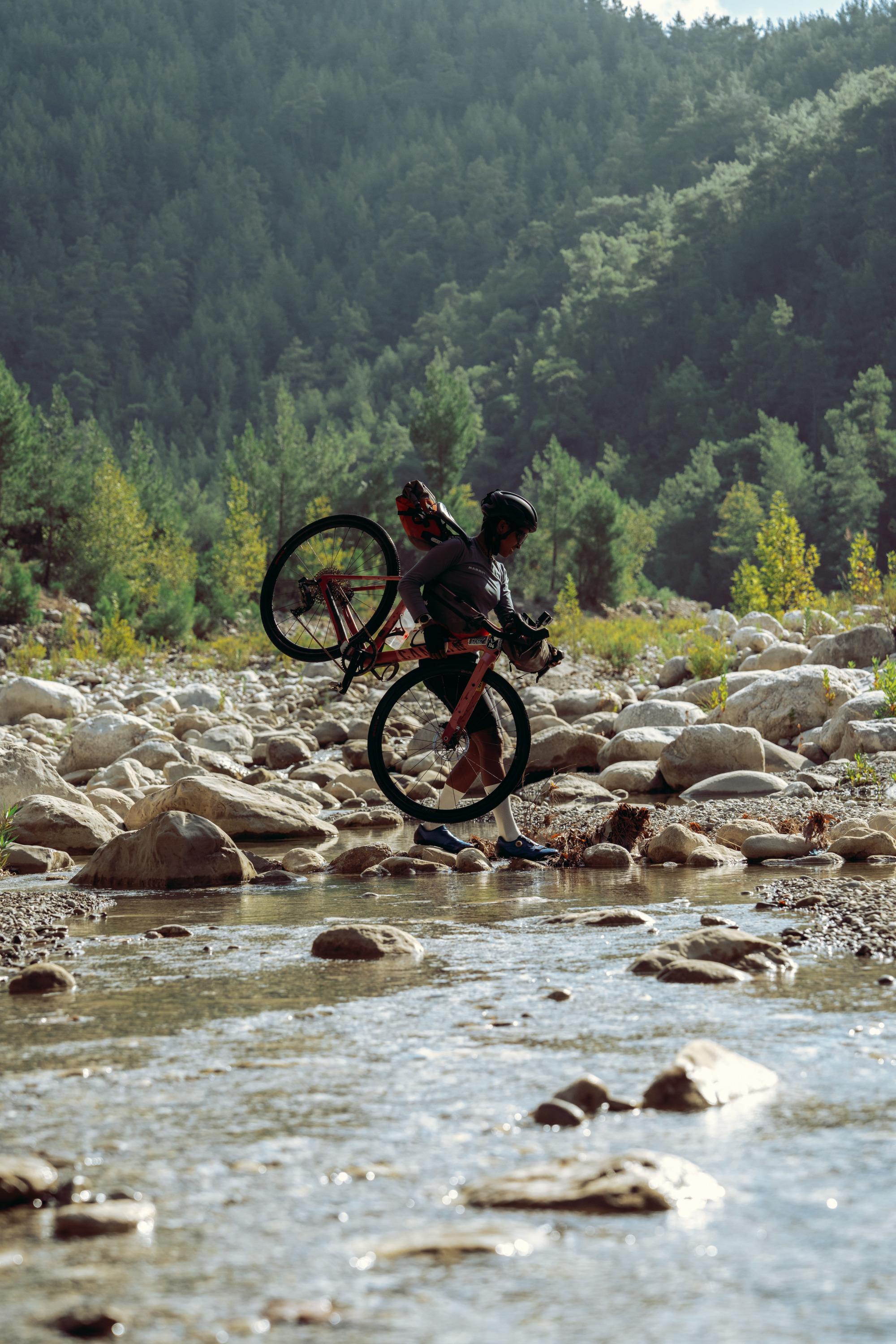 Cyclist carrying a Mammut bike over a rocky stream with forested hills in the background, showcasing rugged outdoor adventure.