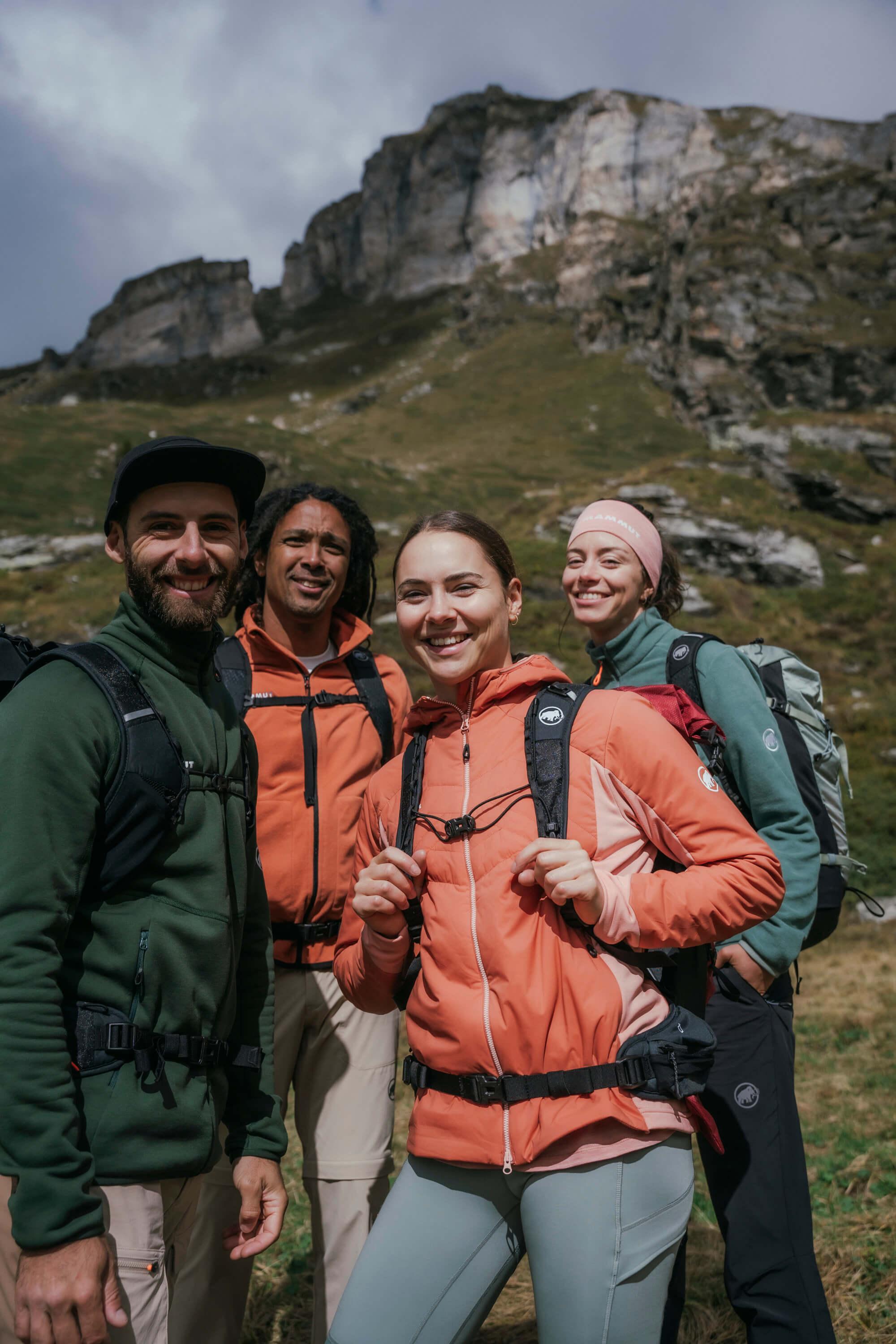 Four Mammut-equipped hikers in high-performance outdoor gear smile at the camera with a majestic mountainous landscape in the background.