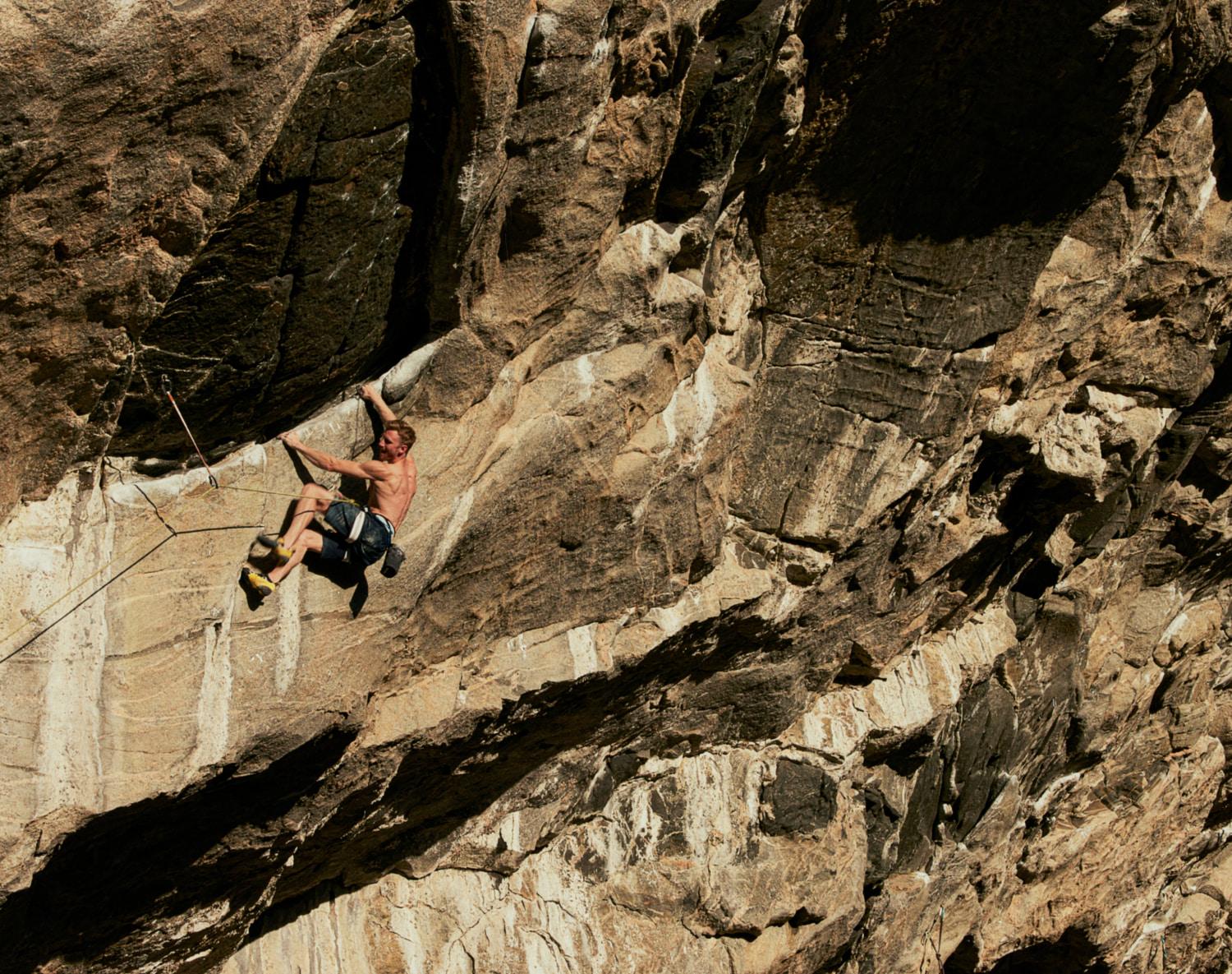 A shirtless climber in Mammut shorts scales a rugged, steep rock face with safety ropes attached, showcasing the brand's high-performance gear for extreme sports and mountaineering.