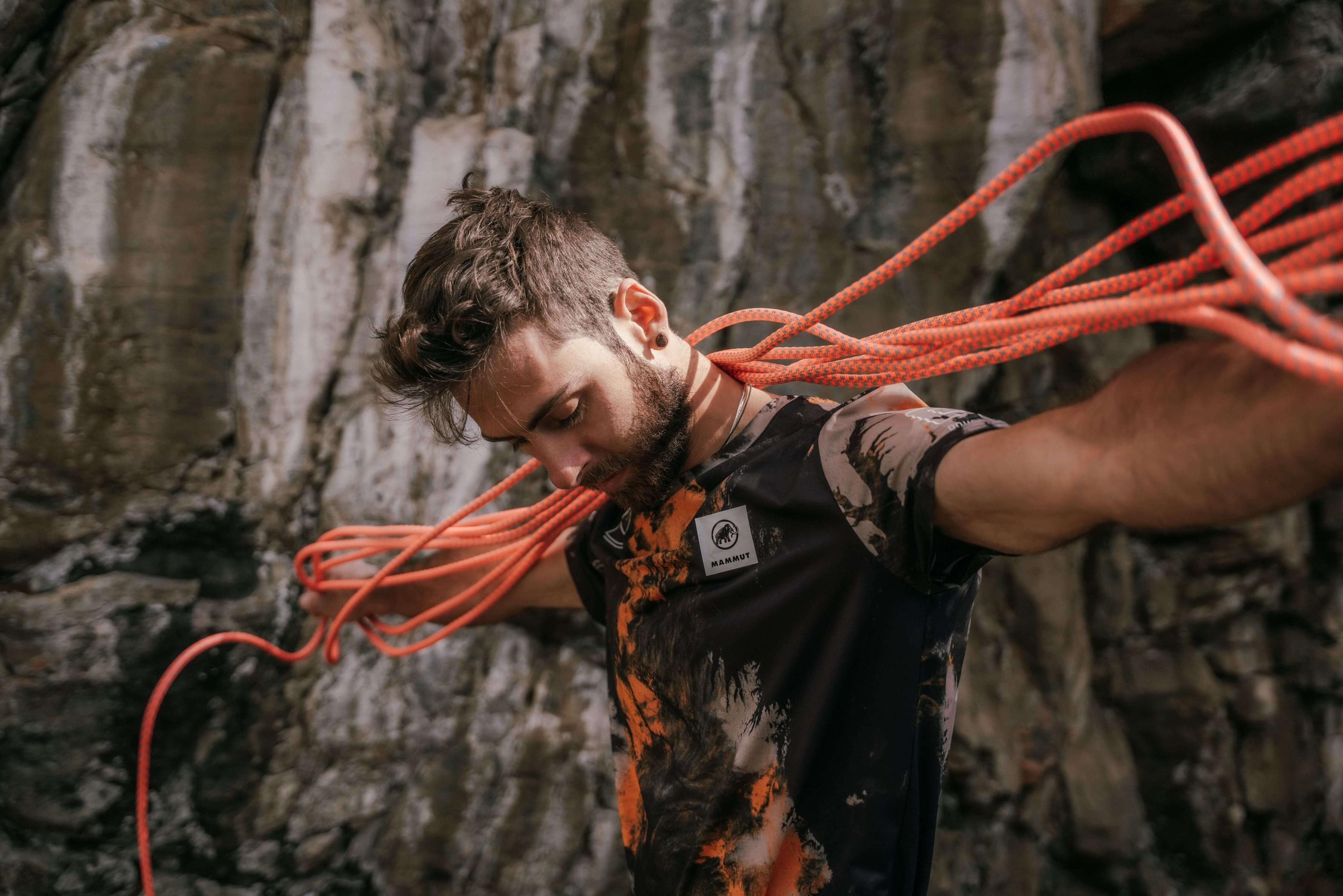 A climber wearing Mammut gear, standing in front of a rocky cliff, drapes orange climbing ropes over his shoulders while looking down.