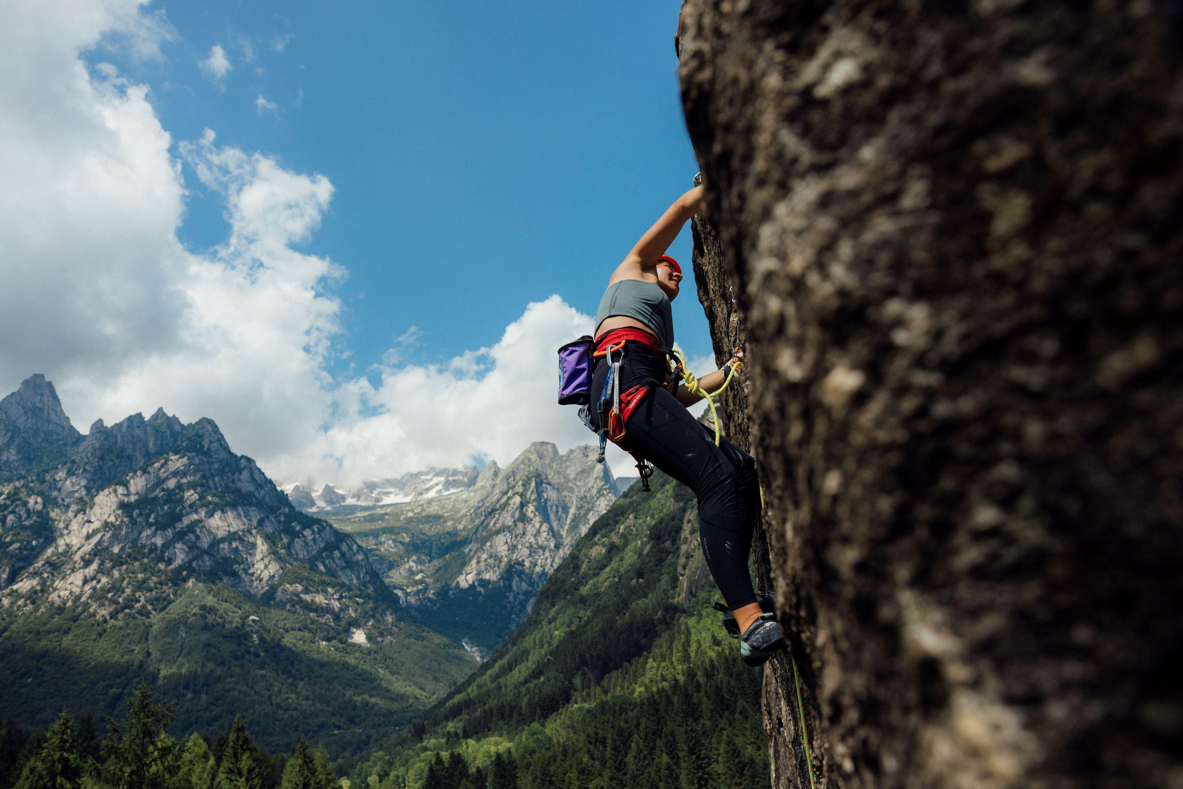 Person rock climbing on a steep cliff face with Mammut gear, set against a backdrop of towering mountains and lush green foliage.