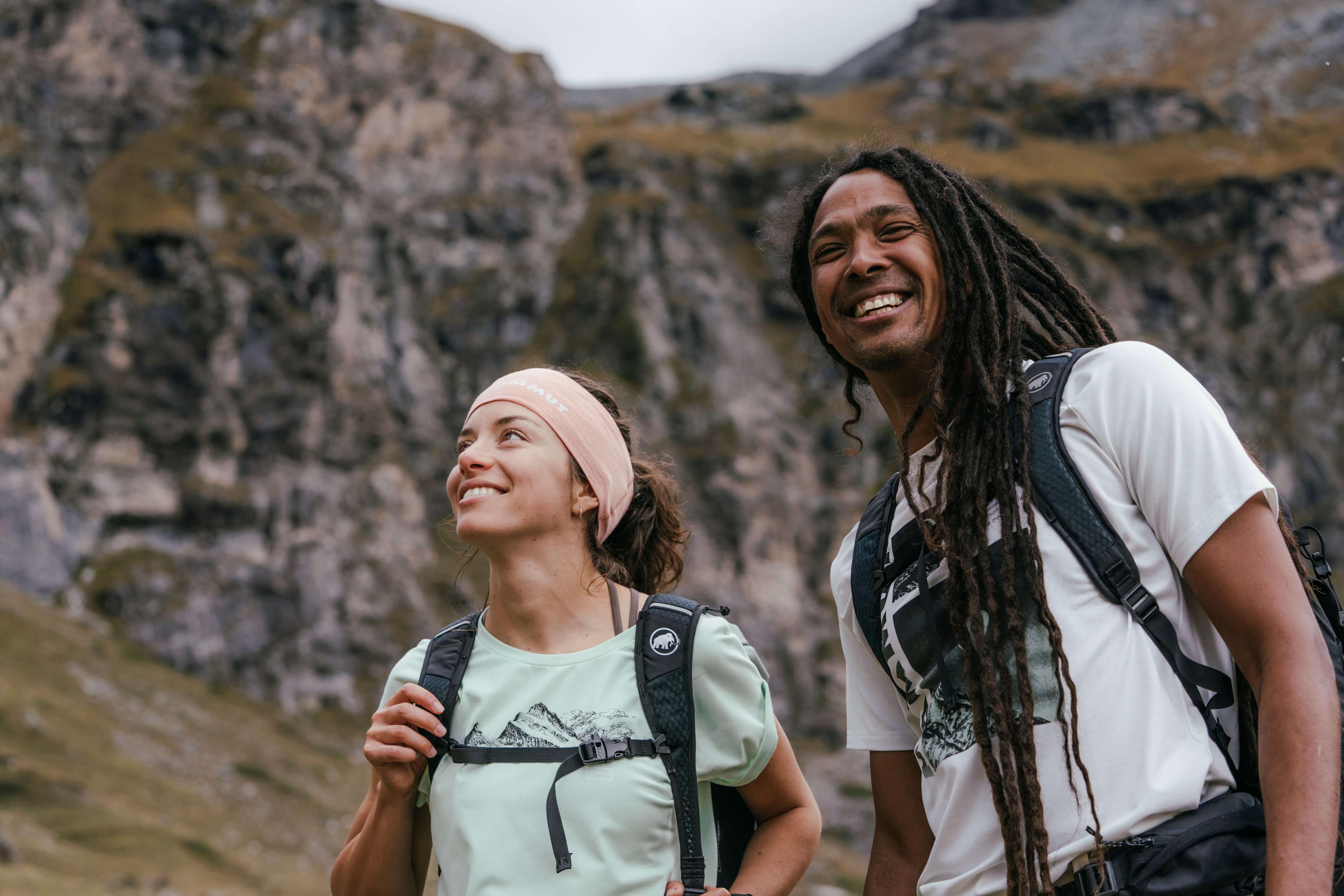 Two hikers wearing Mammut backpacks, smiling and looking up, stand in front of a rugged rocky mountain landscape.