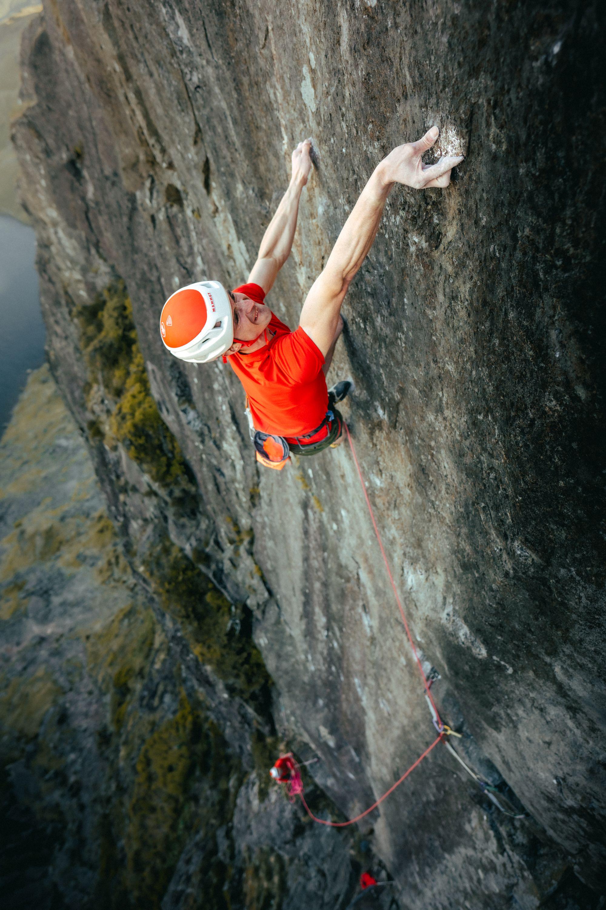 A climber in a red Mammut shirt and helmet scales a steep rocky cliff, secured by a climbing rope.