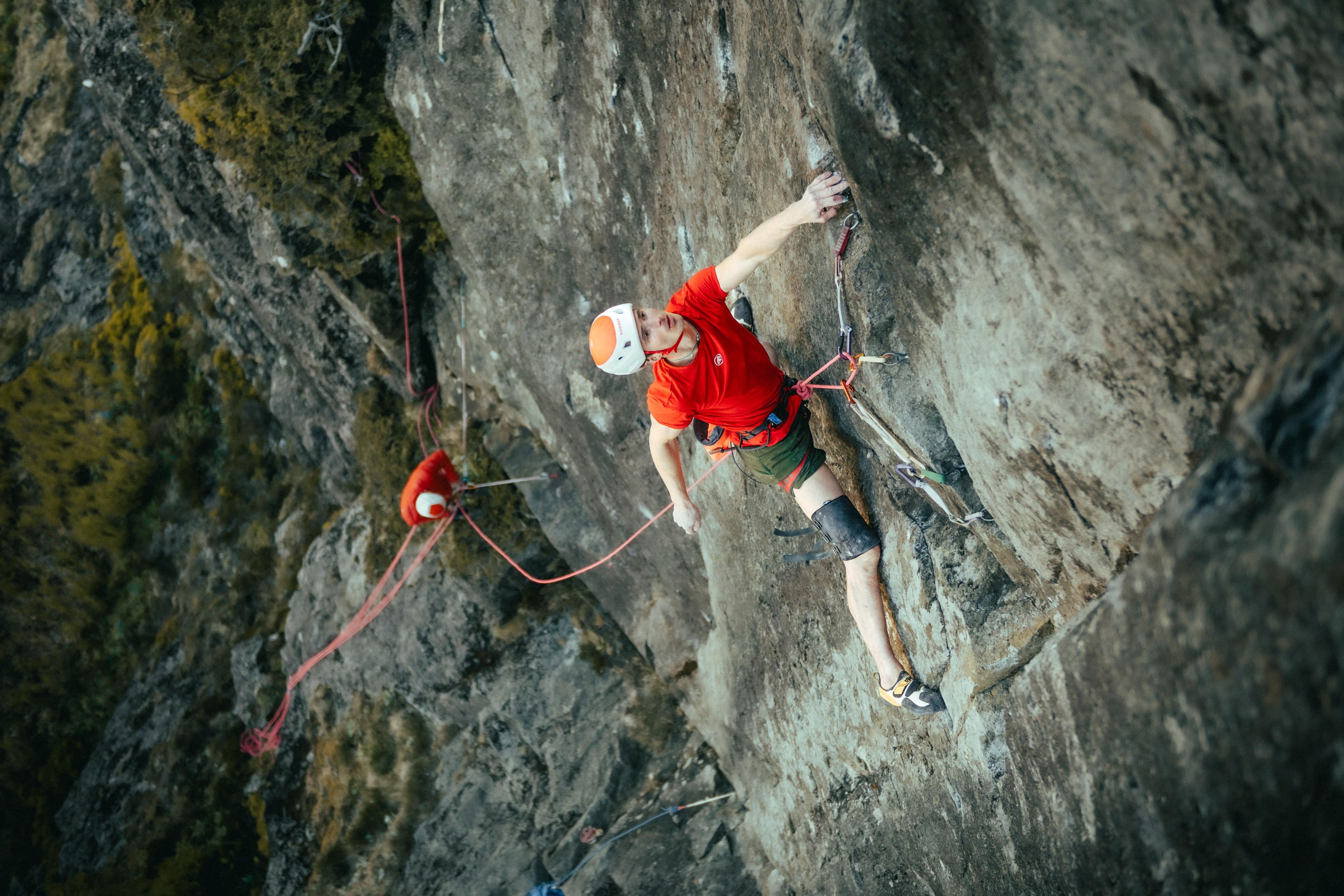 A climber in a red shirt, wearing Mammut climbing gear, ascends a steep rocky cliff using ropes and safety equipment.