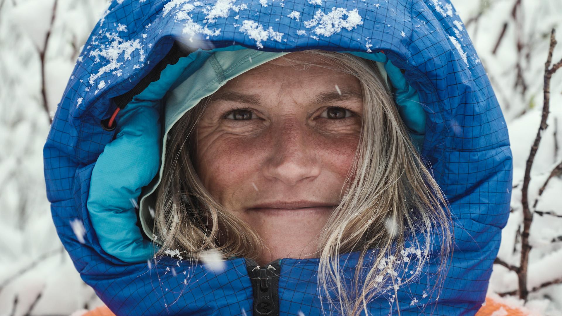 A smiling person wearing a blue Mammut hooded winter jacket, with snowflakes on their face and hood.