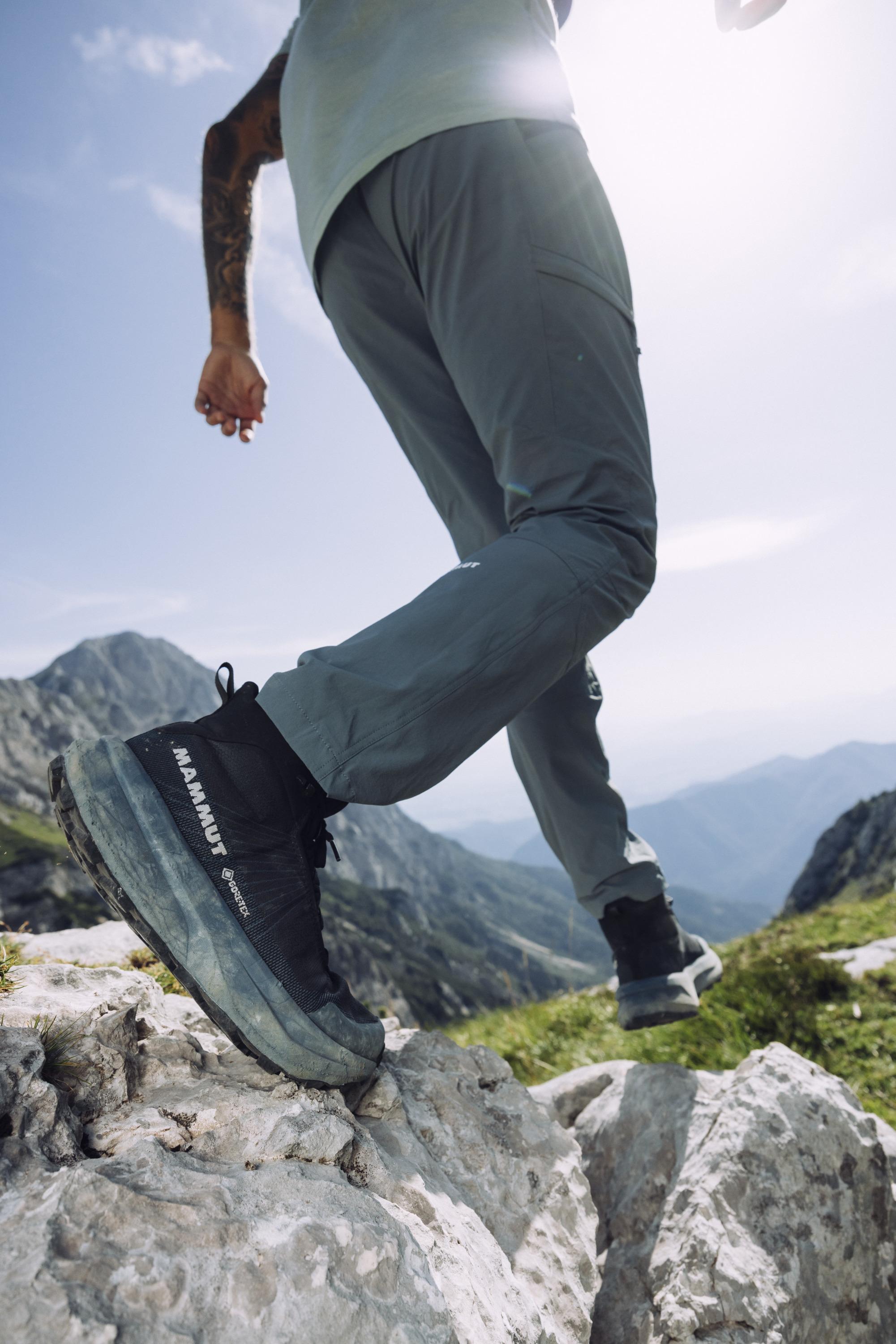 Person hiking on rocky terrain, wearing Mammut gray pants and black shoes, with majestic mountain range in the background.