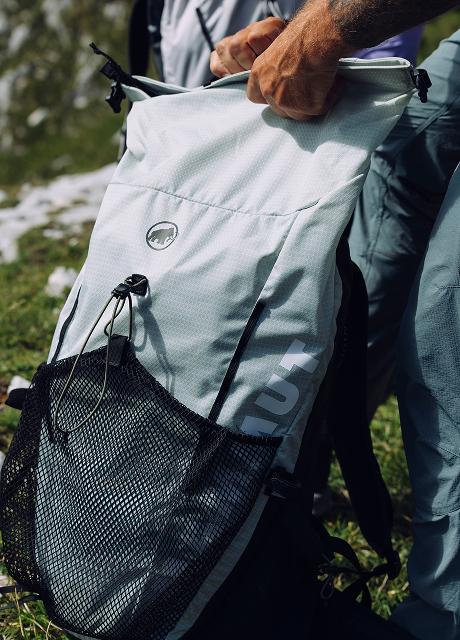Close-up of a person’s hand opening a Mammut light gray hiking backpack with a mesh pocket, outdoors on grass.