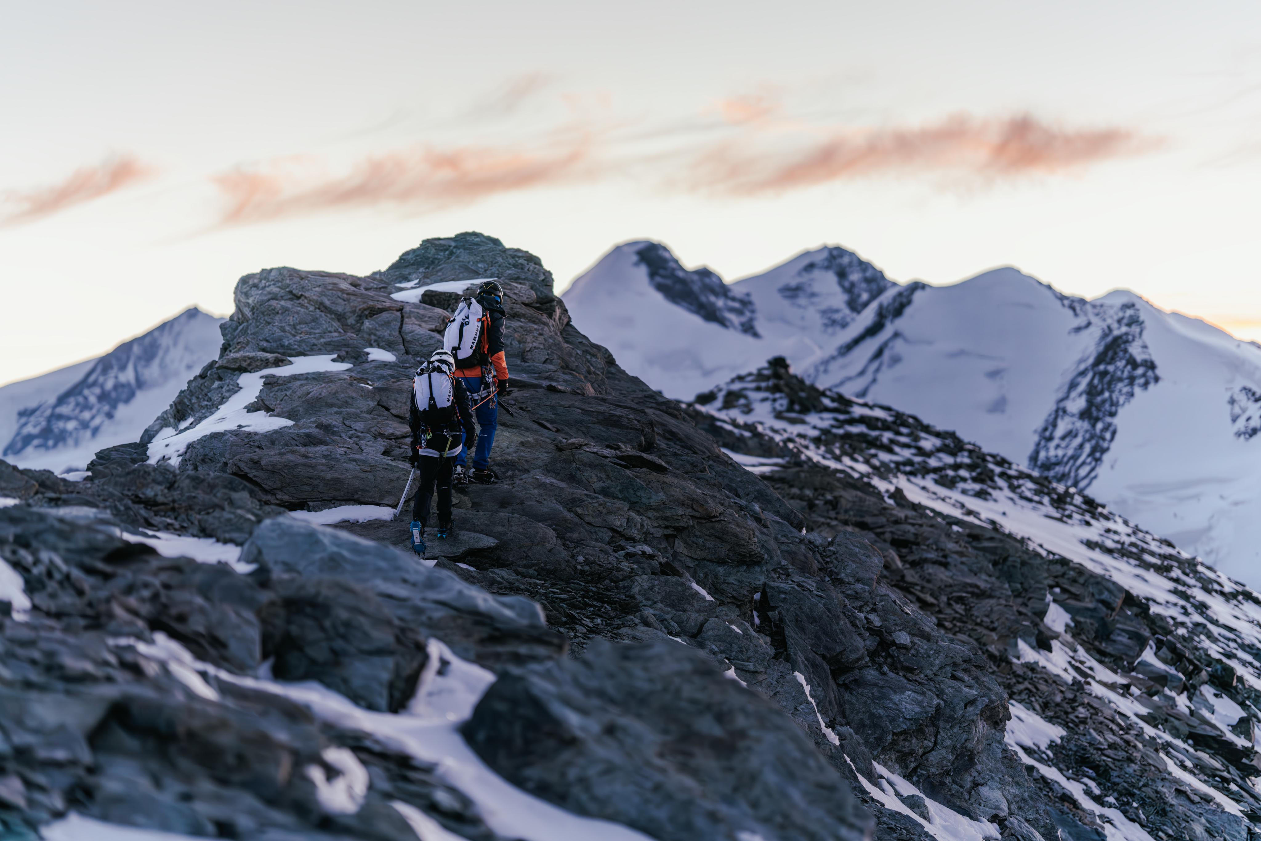 Two climbers with Mammut backpacks traverse a rocky mountain ridge, surrounded by snow-covered peaks at dawn.