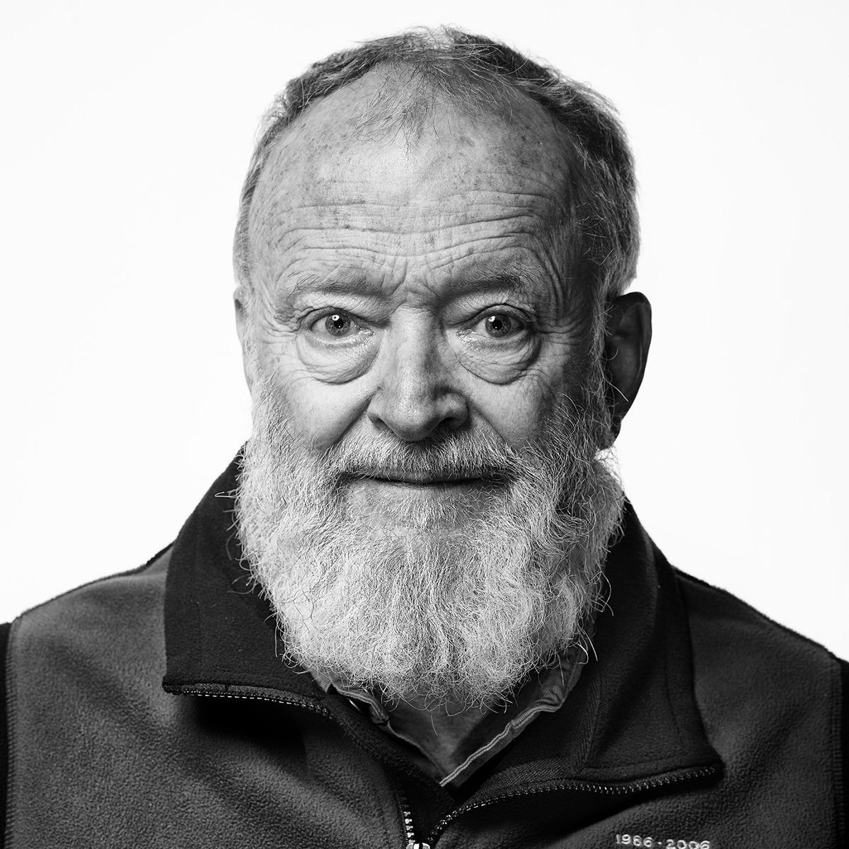 A black and white portrait of an older man with a full white beard, wearing a zipped-up Mammut mountaineering jacket.