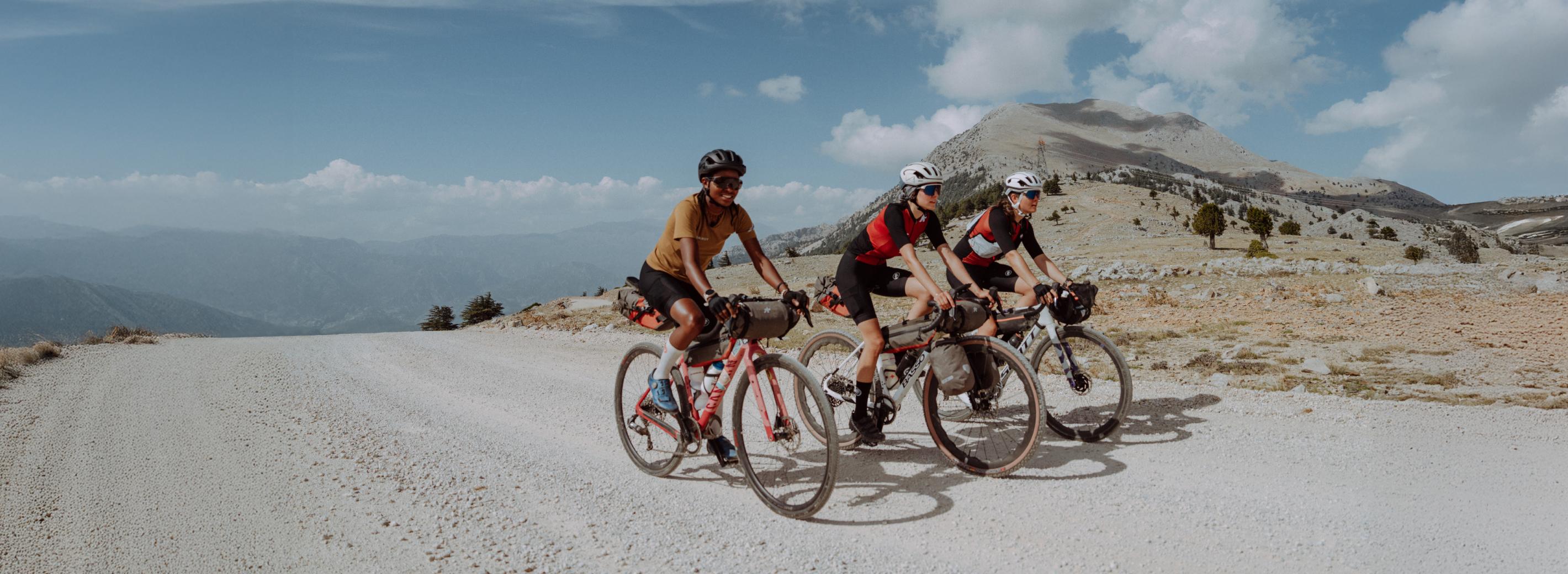 Three cyclists wearing Mammut gear ride on a scenic gravel road, framed by majestic mountains and a partly cloudy sky in the background.