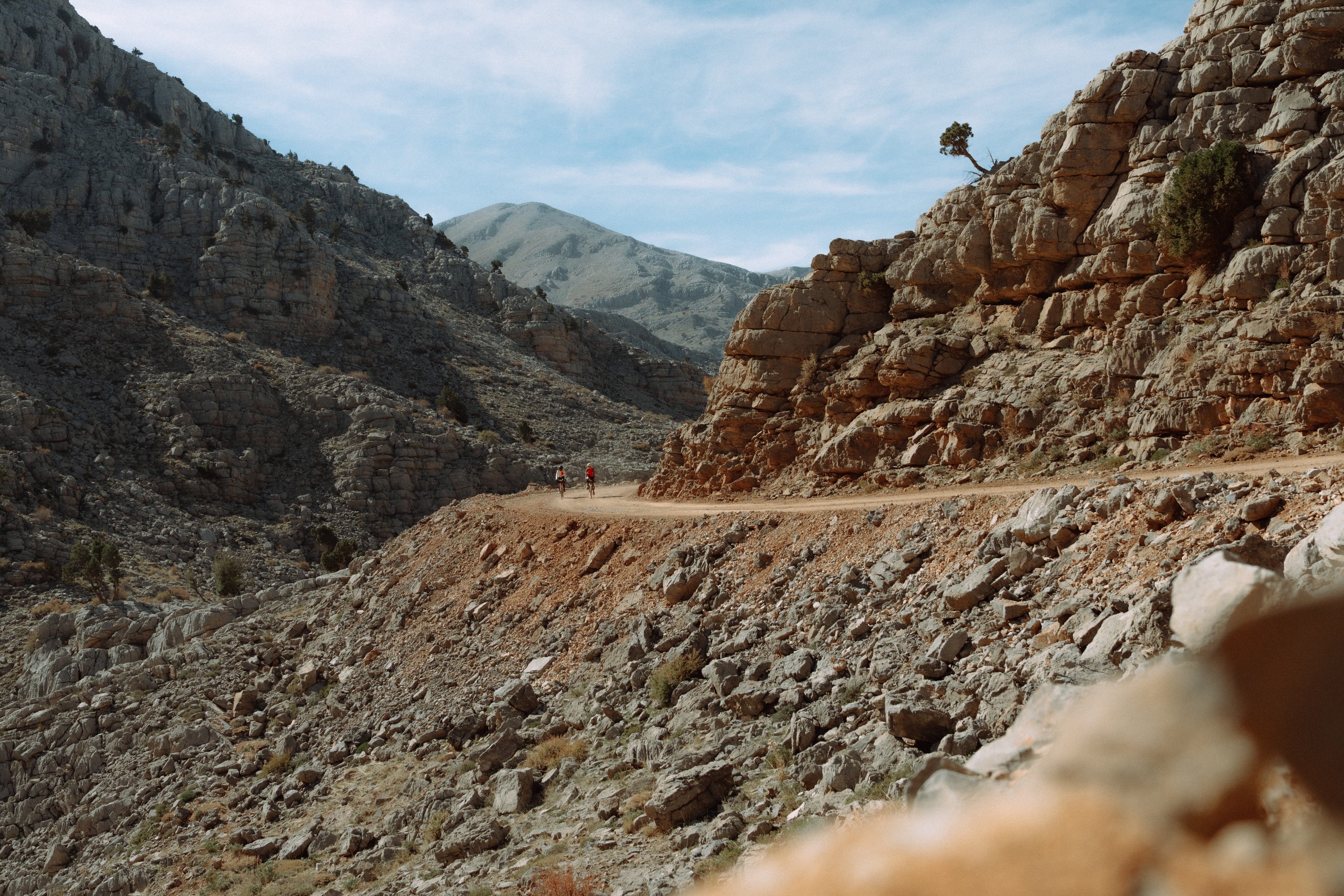 Rugged mountain trail winding through a rocky landscape, featuring distant hikers sporting Mammut gear under a clear blue sky.