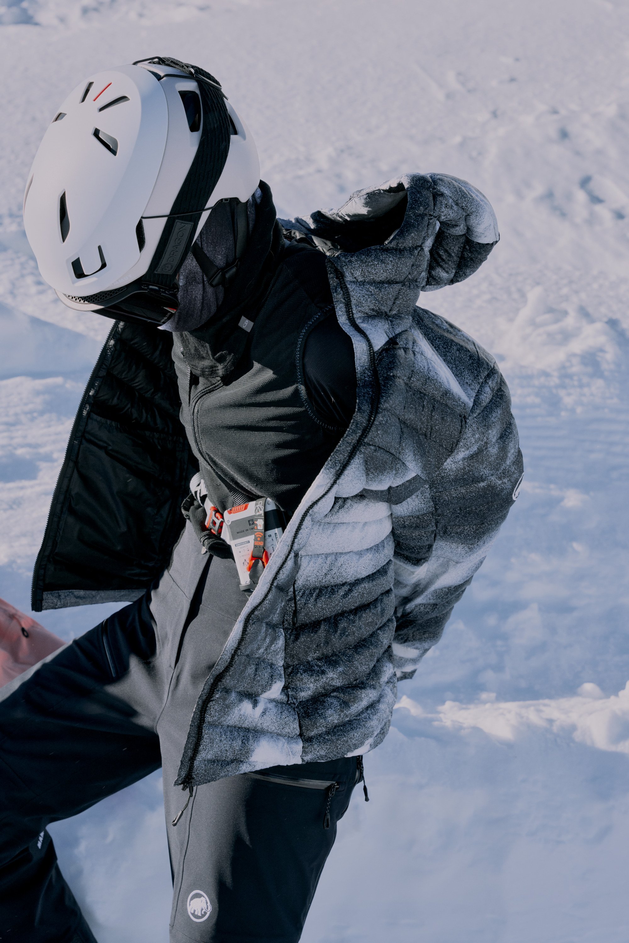 A hiker with a headlamp on a trail at dusk, equipped with Mammut winter clothing and backpack, against a backdrop of a snowy mountain.