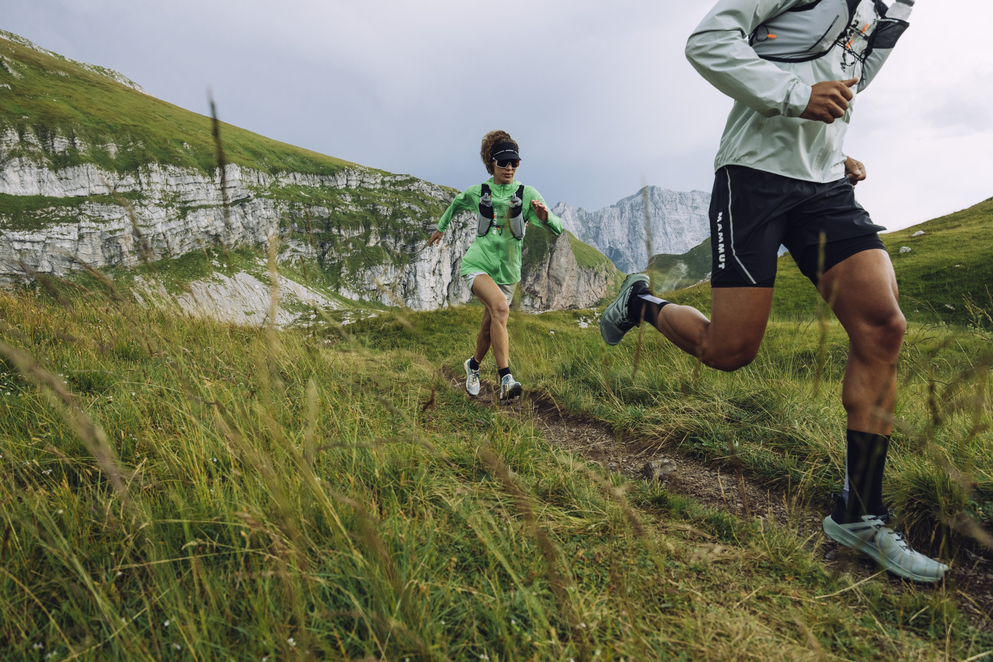 Two people trail running on a grassy mountain path with rocky cliffs in the background, wearing Mammut outdoor gear.