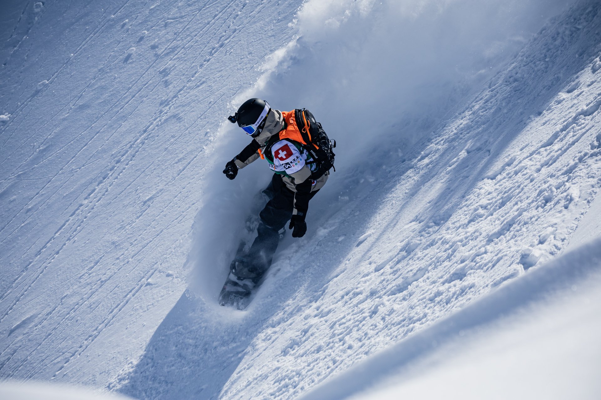 A Mammut skier in mid-air performing an impressive jump off a snowy cliff with stunning snow-covered mountains in the background.