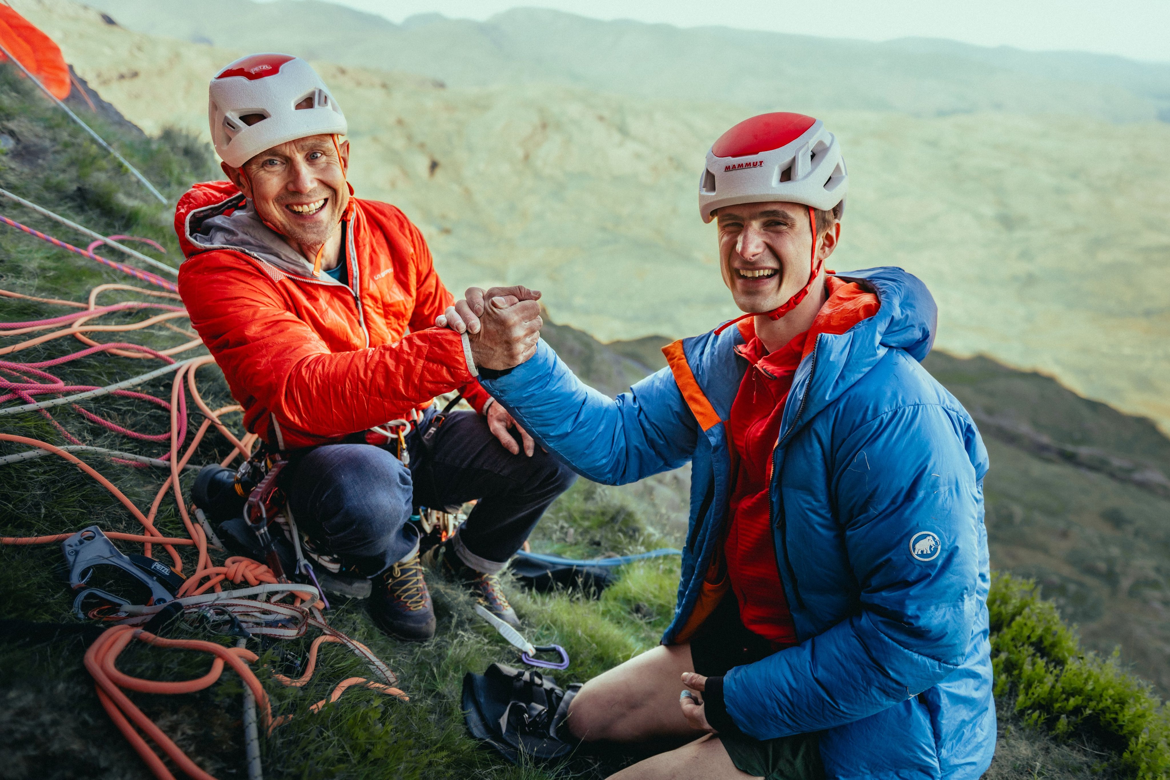 Two climbers wearing helmets and bright Mammut jackets smile and fist bump on a rocky mountainside, equipped with Mammut climbing gear.