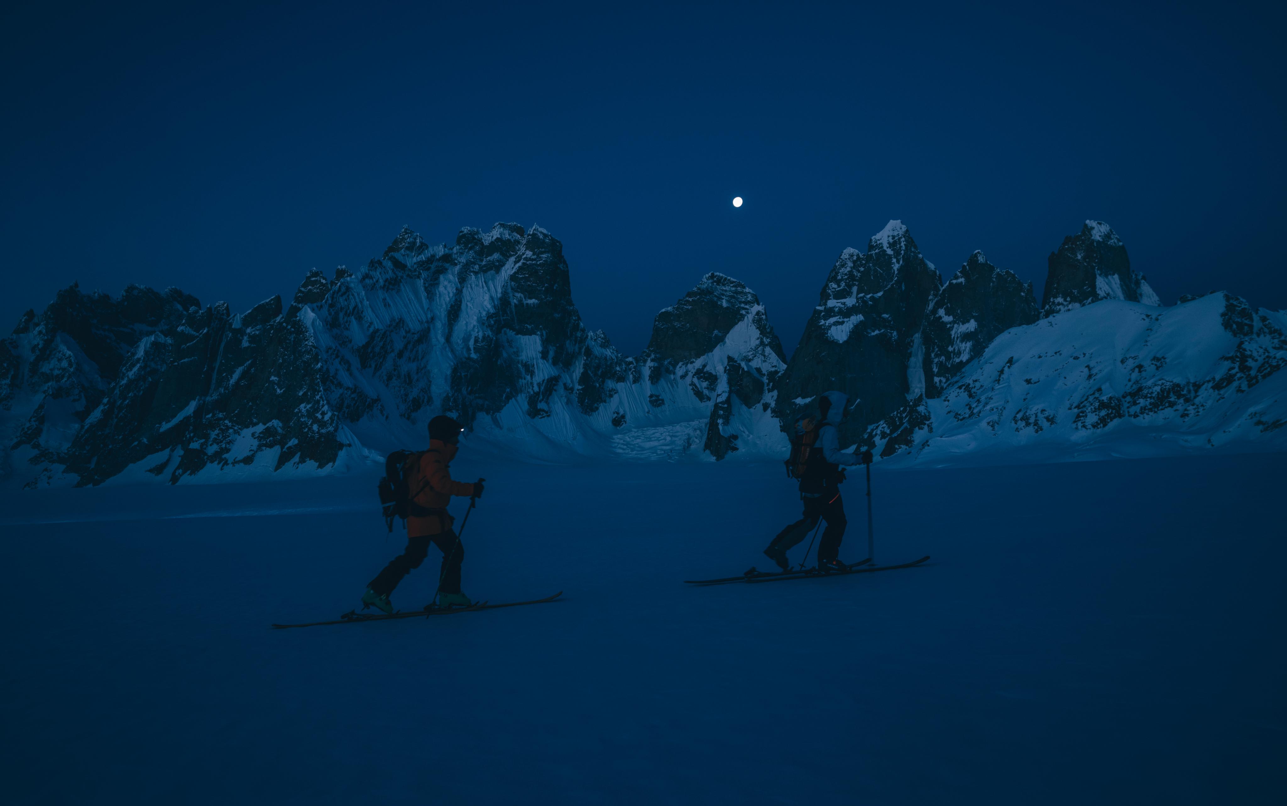 Two skiers in Mammut gear glide under a bright moonlit sky through a pristine, snow-covered mountainous landscape.