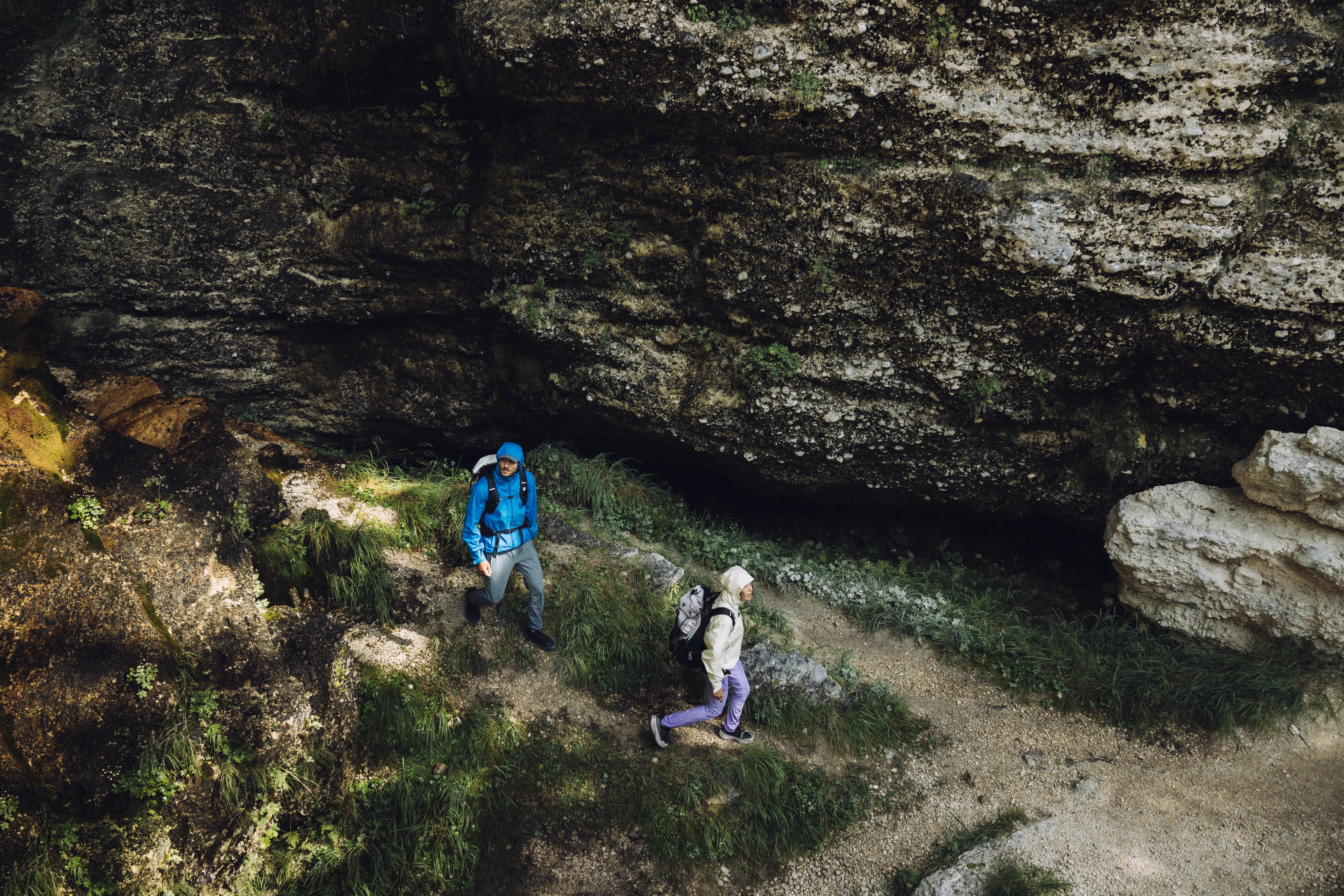 Four hikers wearing Mammut backpacks and outdoor gear climb a rocky, mountainous trail under a cloudy sky.