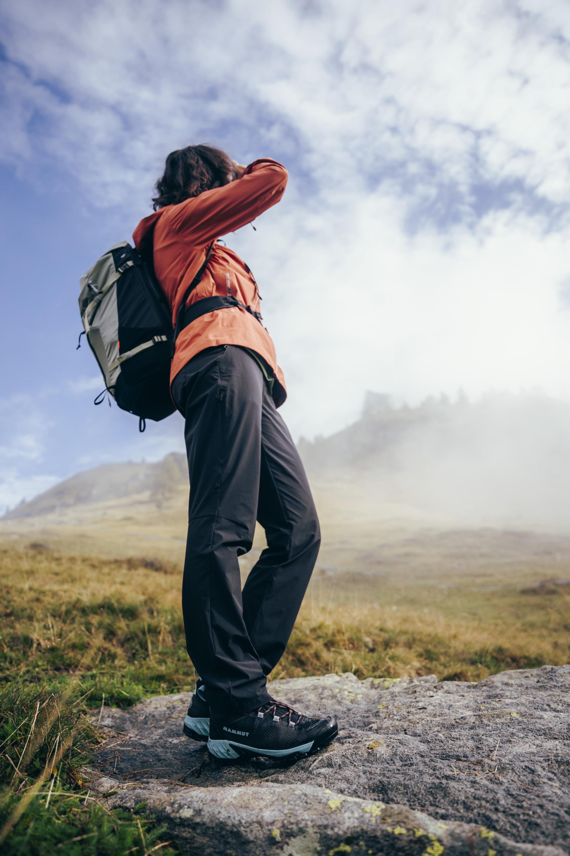 Person wearing an orange Mammut jacket and black pants, carrying a backpack, standing on a rock, gazing at a foggy mountain landscape.