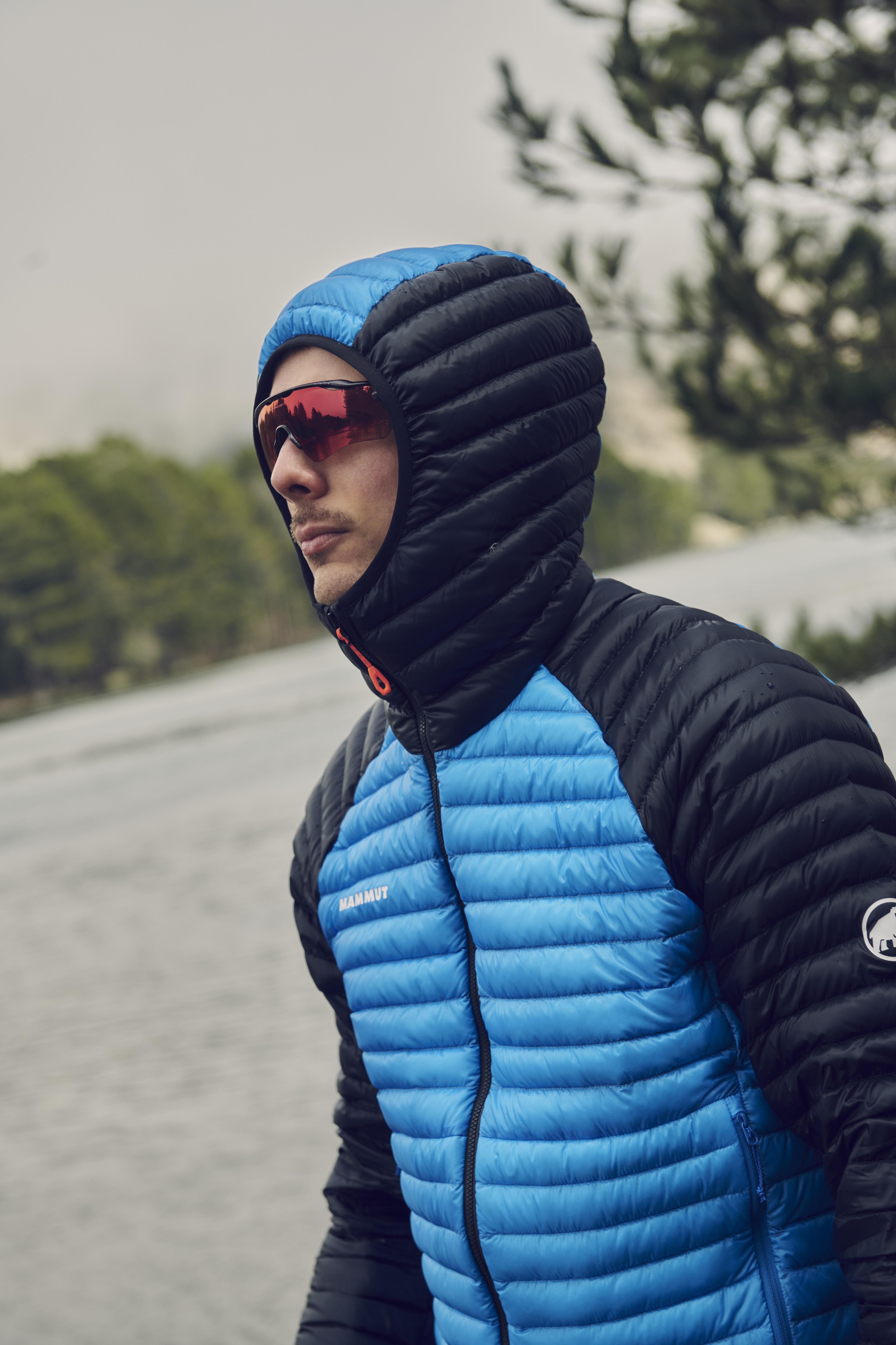 Individual dressed in a purple Mammut puffer jacket with the hood up, standing outside in a mountainous landscape.