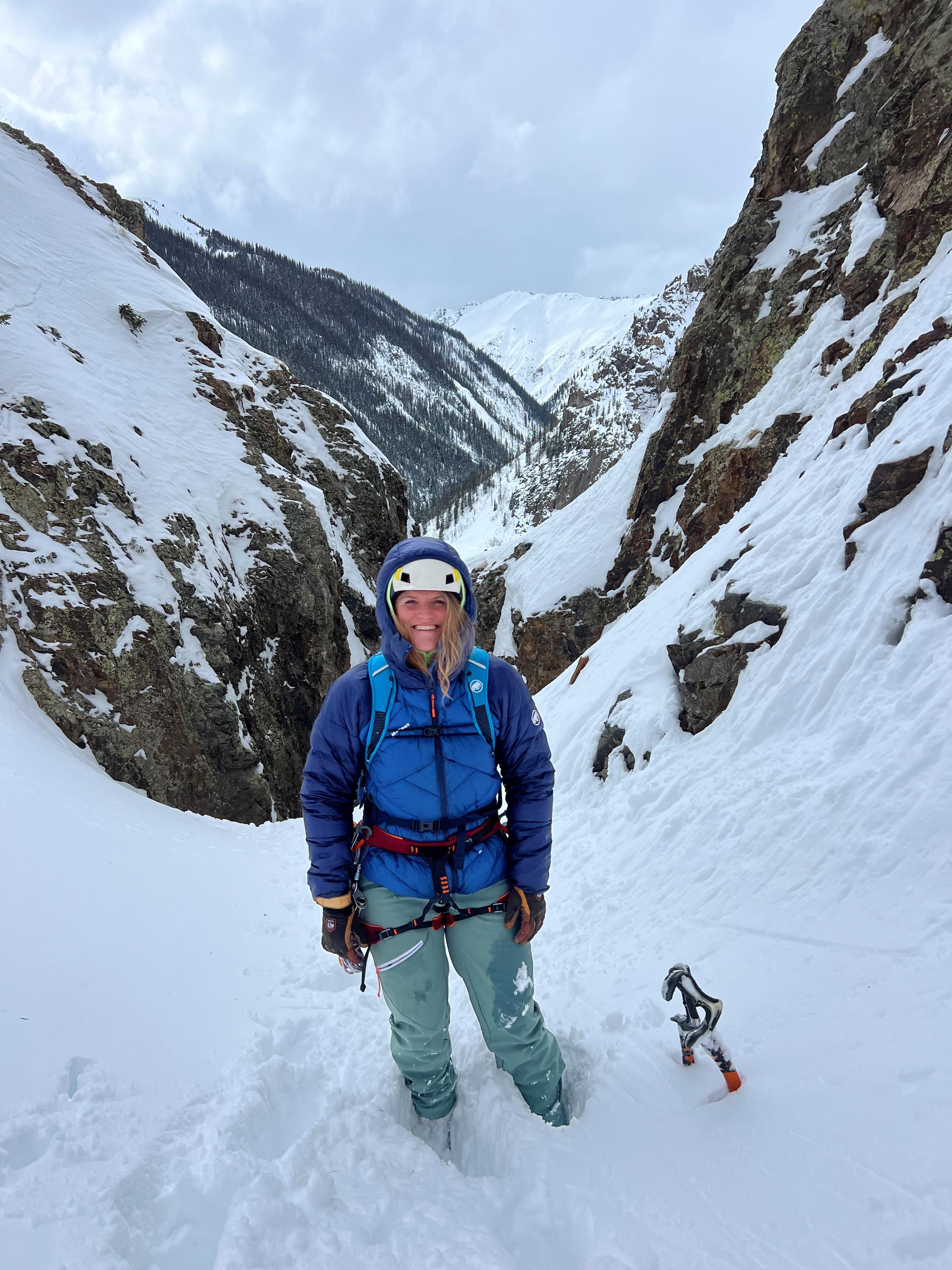 Mountaineer in Mammut winter gear stands in a snowy mountain pass with an ice axe nearby.