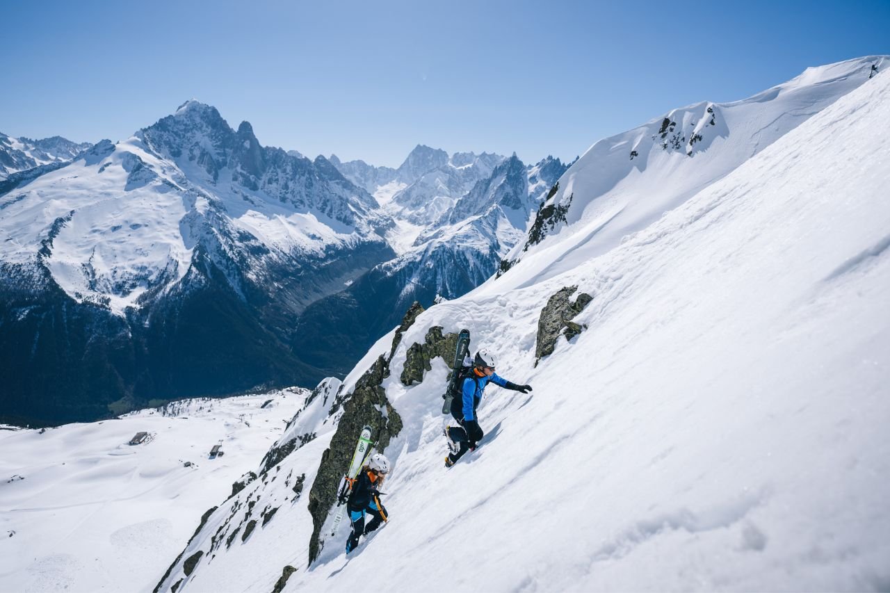 Two climbers wearing Mammut gear ascend a steep, snowy mountain slope with snow-covered peaks in the background under a clear sky.
