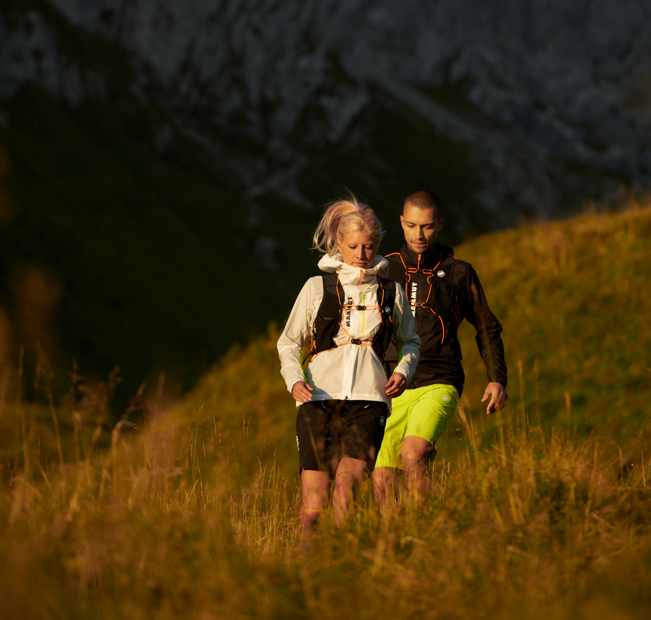 Two hikers wearing Mammut backpacks and outdoor gear trekking a sunny mountain trail.