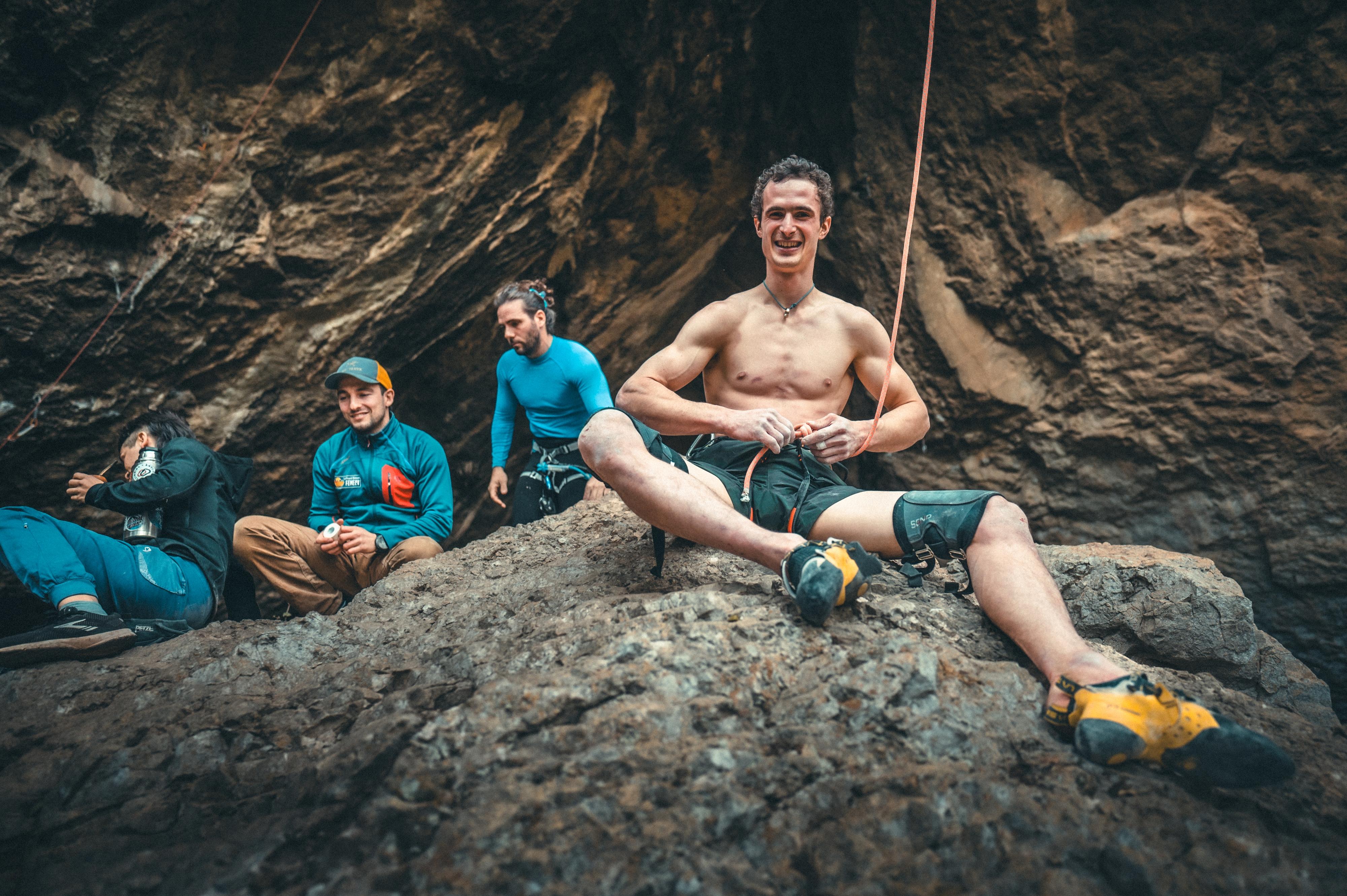 Shirtless man smiles at camera while sitting on rocks, wearing Mammut climbing gear; three people in the background with Mammut equipment near a cave wall.
