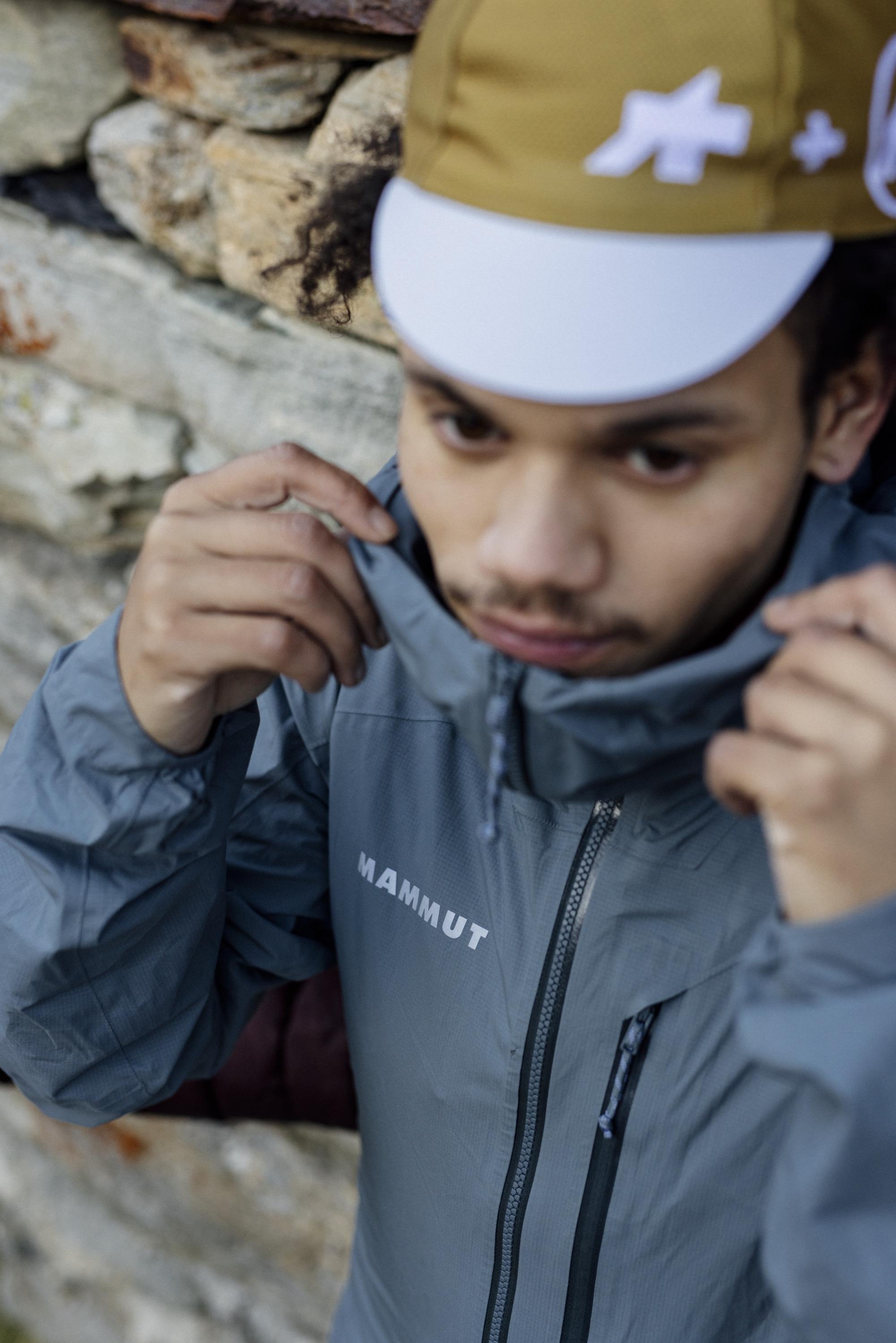 Person wearing a gray Mammut outdoor jacket and a yellow cap near a stone wall, adjusting the collar for optimal fit.