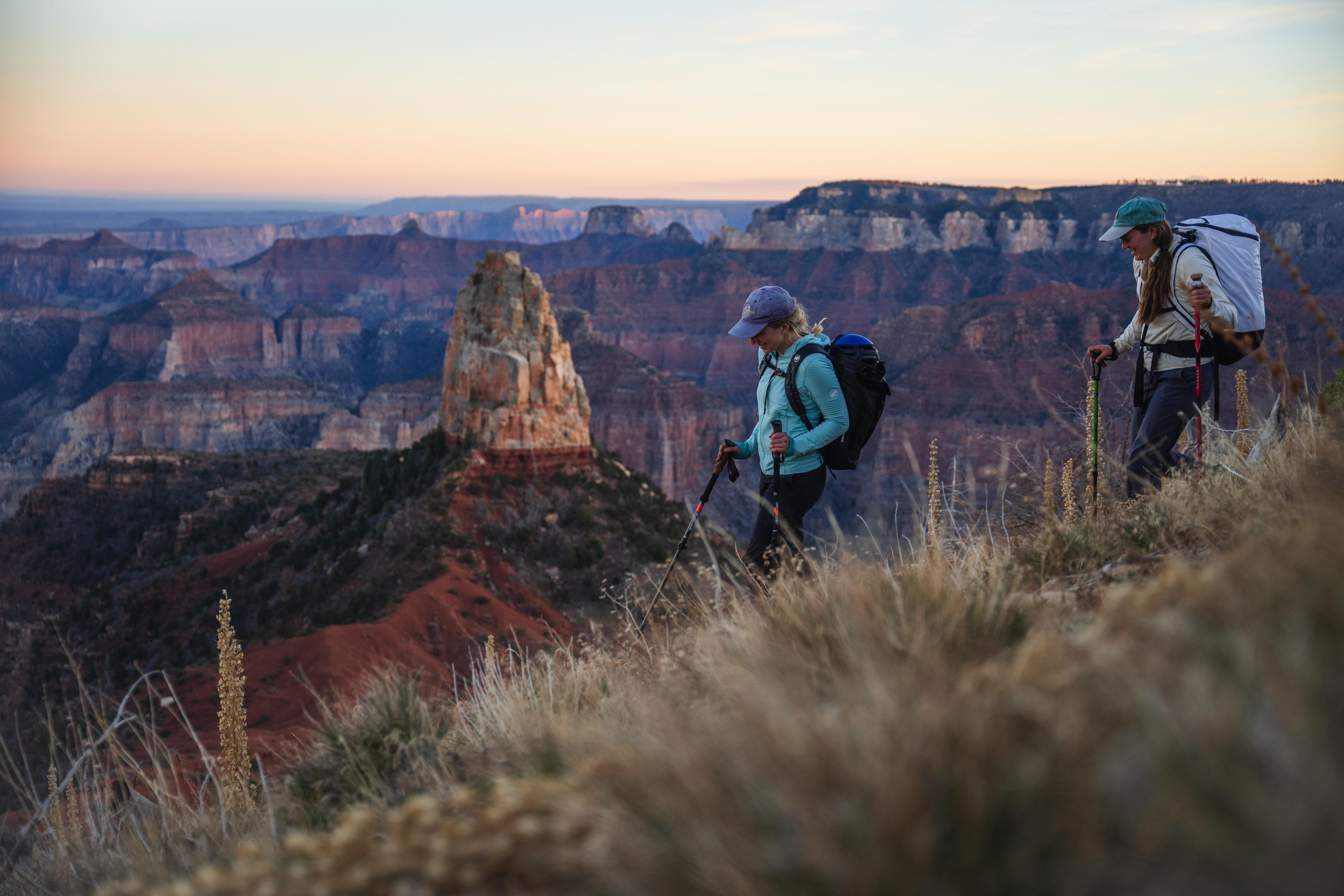 Two Mammut-equipped hikers with backpacks and hiking poles walking along a trail, enjoying a stunning sunrise view of a canyon.