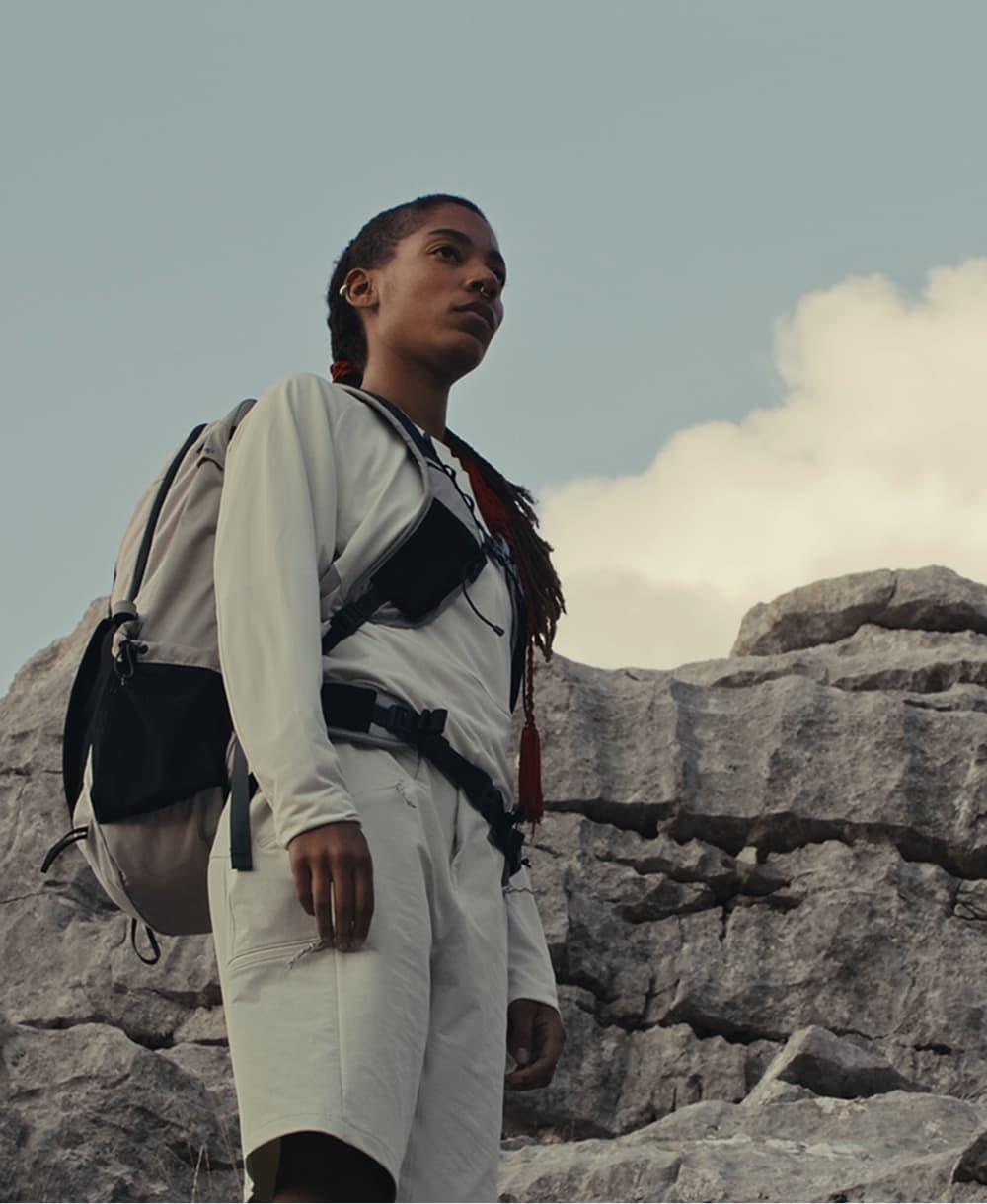 Person in white Mammut outdoor gear stands on rocky terrain, gazing upward, with alpine mountains and clear blue sky behind.